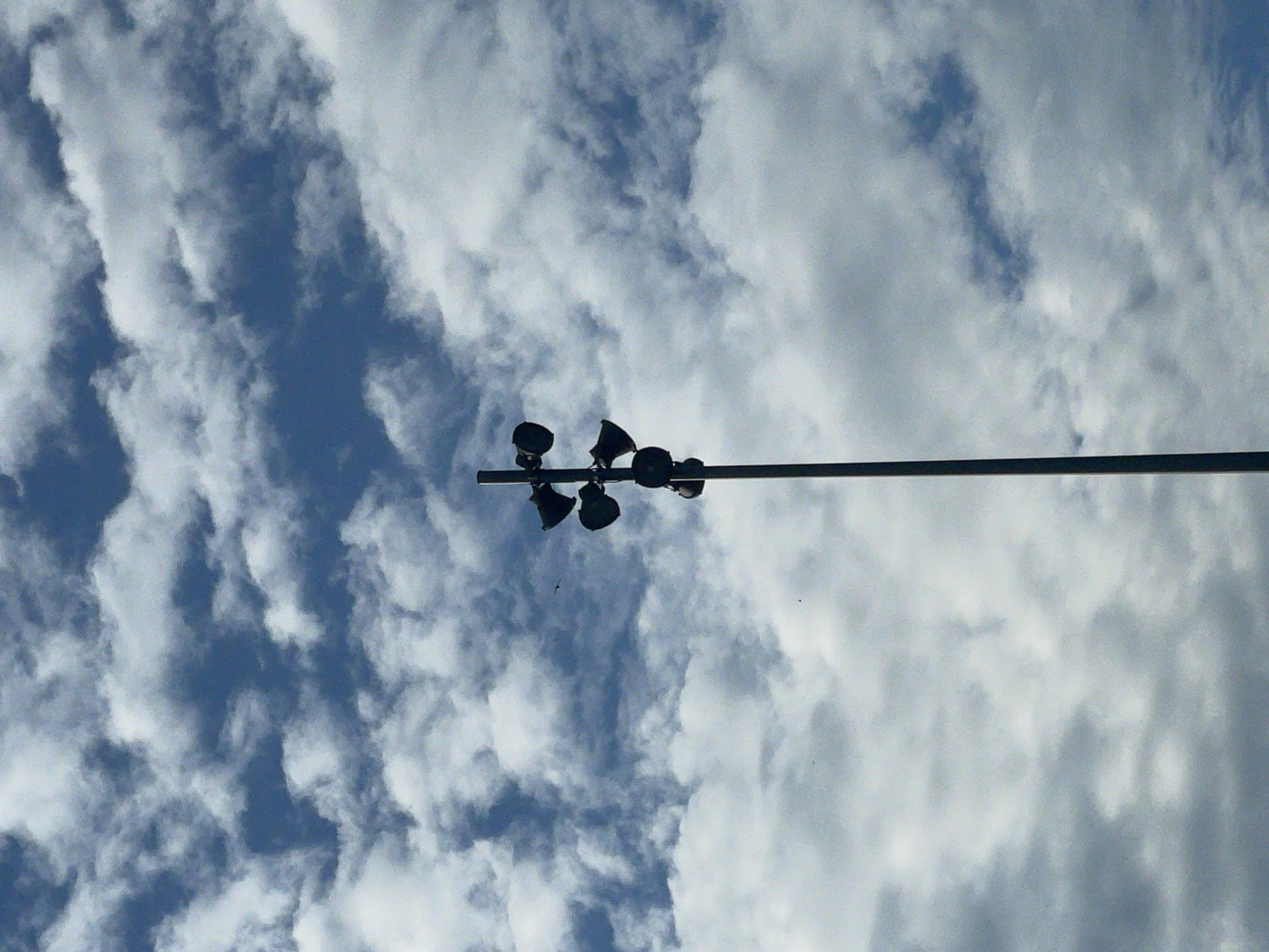 The image is a low-angle shot of a metal pole supporting several lights. The lights are dark and silhouetted against a cloudy sky. The sky transitions from deep blue to pale gray-white with textured cloud formations. The photo was taken in Mykolaiv, Ukraine. No people are visible.