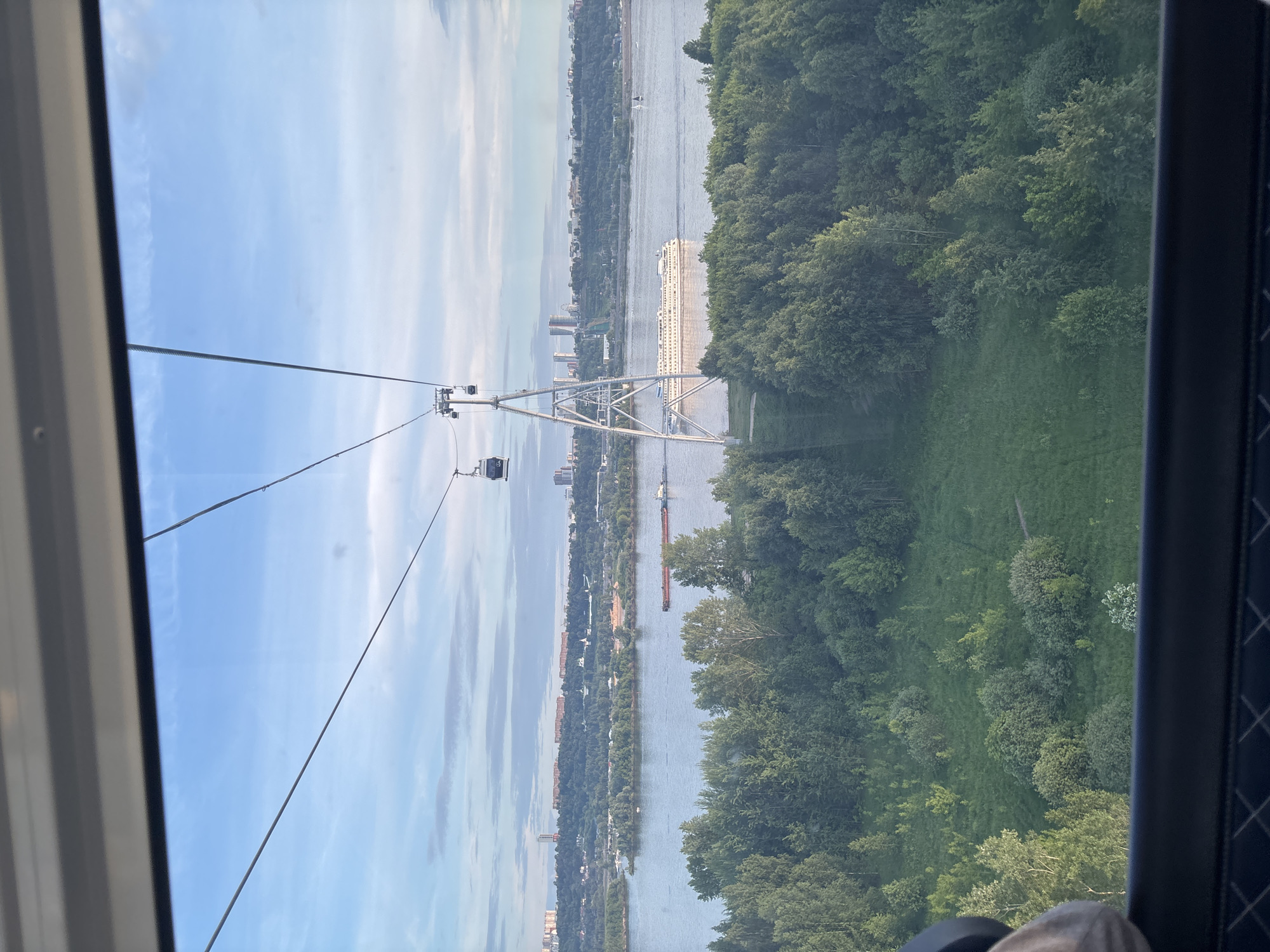 This image captures a panoramic view from a cable car crossing a wide river, likely the Volga River near Bor, Russia. The perspective is from inside a cable car cabin, with parts of the window frame visible on the left and right edges. In the bottom right corner, a partial view of a person's head or hat is visible, suggesting a passenger.
The scene features a prominent metal pylon (cable car tower) supporting numerous cables that stretch across the river. A cable car gondola is visible in the distance, further along the line. The river itself dominates the midground, with a large, white multi-deck passenger cruise ship sailing on its surface, moving from left to right. Another, smaller, darker barge-like vessel is visible further downstream.
On the left bank of the river, a cityscape or town is spread out, featuring various buildings, some taller structures, and green foliage along the river's edge. The right bank is characterized by dense, verdant forest, with a grassy area or hillside visible beneath the trees, directly below the cable car's path.
The time of day appears to be bright daylight, possibly morning or afternoon, indicated by the clear blue sky with scattered white clouds and good visibility. The weather seems fair, contributing to the scenic quality of the elevated view. No specific text is discernible within the image.