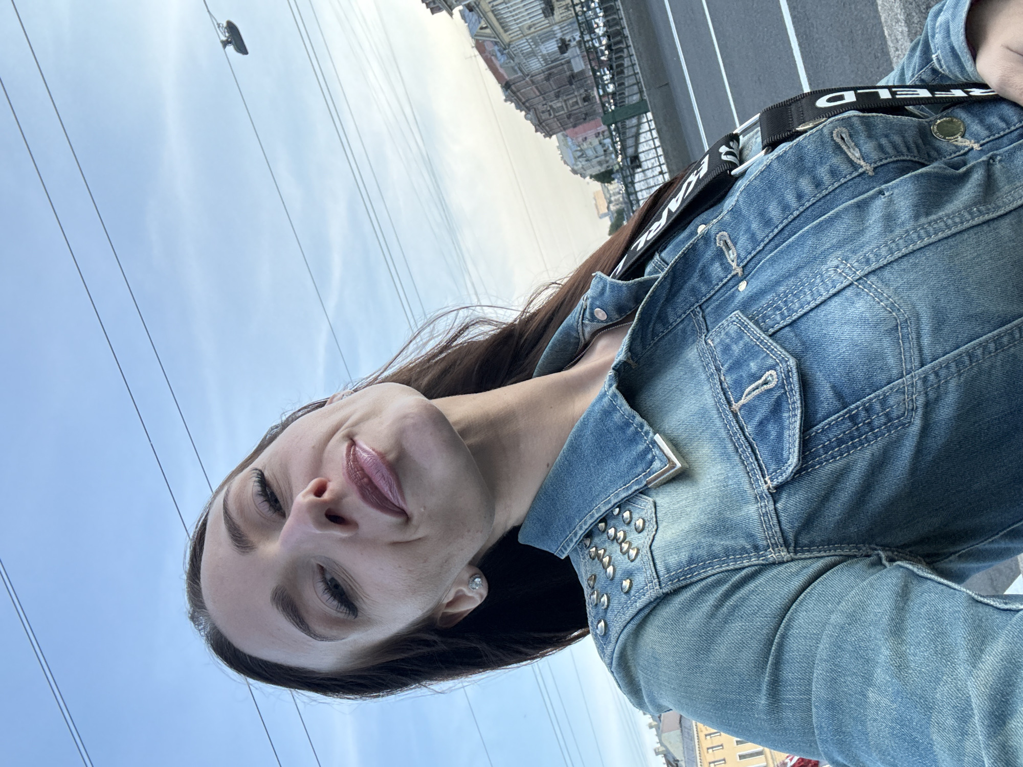 A woman takes a selfie outdoors, looking up towards the sky. She has long, dark hair and is wearing a blue denim jacket adorned with silver studs on the collar and across the chest. A black strap with white lettering is visible across her chest. The background shows a bright sky with some clouds and electrical wires. In the upper portion of the image, buildings typical of Saint Petersburg, Russia, can be seen, suggesting an urban setting. The lighting indicates it is daytime, likely late afternoon given the soft light. The woman's expression is a slight smile, conveying a relaxed or happy mood. The overall impression is a casual, outdoor snapshot in an urban environment. Visible text on the strap appears to be "KARL".