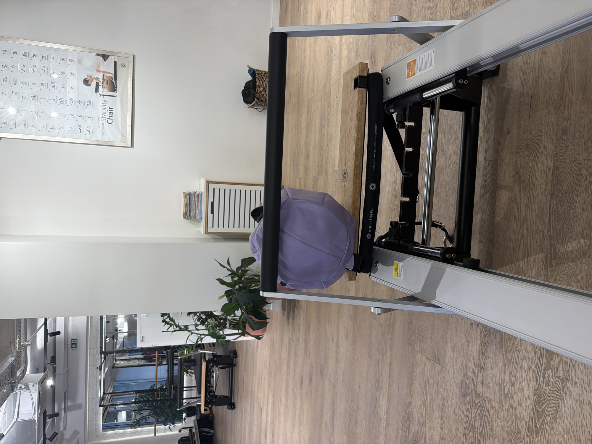 The image depicts a Pilates studio in Boreti, Montenegro. In the foreground, a piece of Pilates equipment, likely a reformer, is prominently displayed. A light purple bag rests on the equipment. The studio features a wooden floor and is illuminated by overhead lighting. In the background, there are large windows revealing an indoor space with gym equipment, plants, and shelving units. A framed poster displaying various Pilates exercises is mounted on the wall. The overall scene suggests a clean and functional fitness environment.