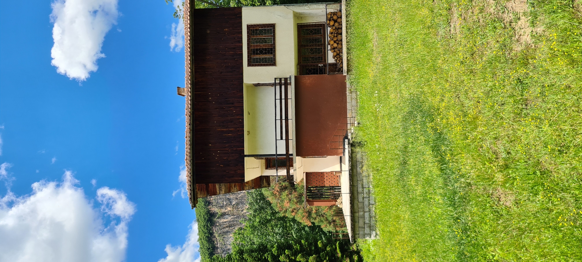 A two-story house with a brown tile roof stands on a slight incline, surrounded by lush green grass and trees. The house has white and brown exterior walls, with large windows and a balcony on the upper floor. In front of the house, a stone foundation supports a concrete patio. To the right of the patio, a stack of cut logs is visible. Behind the house, a steep, rocky cliff face rises, covered in dense green foliage. The sky is a clear blue with scattered white clouds. The scene appears to be a rural residential property in Lopian, Bulgaria, on a sunny day.