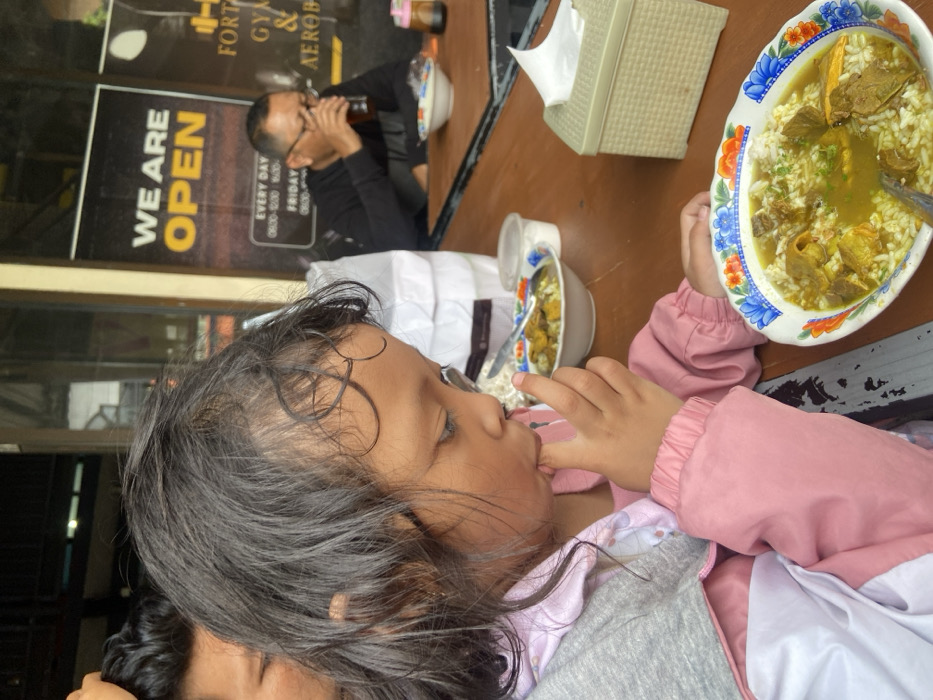 A young child wearing a pink jacket and grey sweater eats with their fingers while seated at a table. Two bowls of soup are visible, one larger with meat, and one smaller. A man drinks from a bottle in the background, sitting next to a sign that says "We Are Open" and advertises a gym and aerobics. A tissue box also sits on the table. The scene appears to be taken outside.
