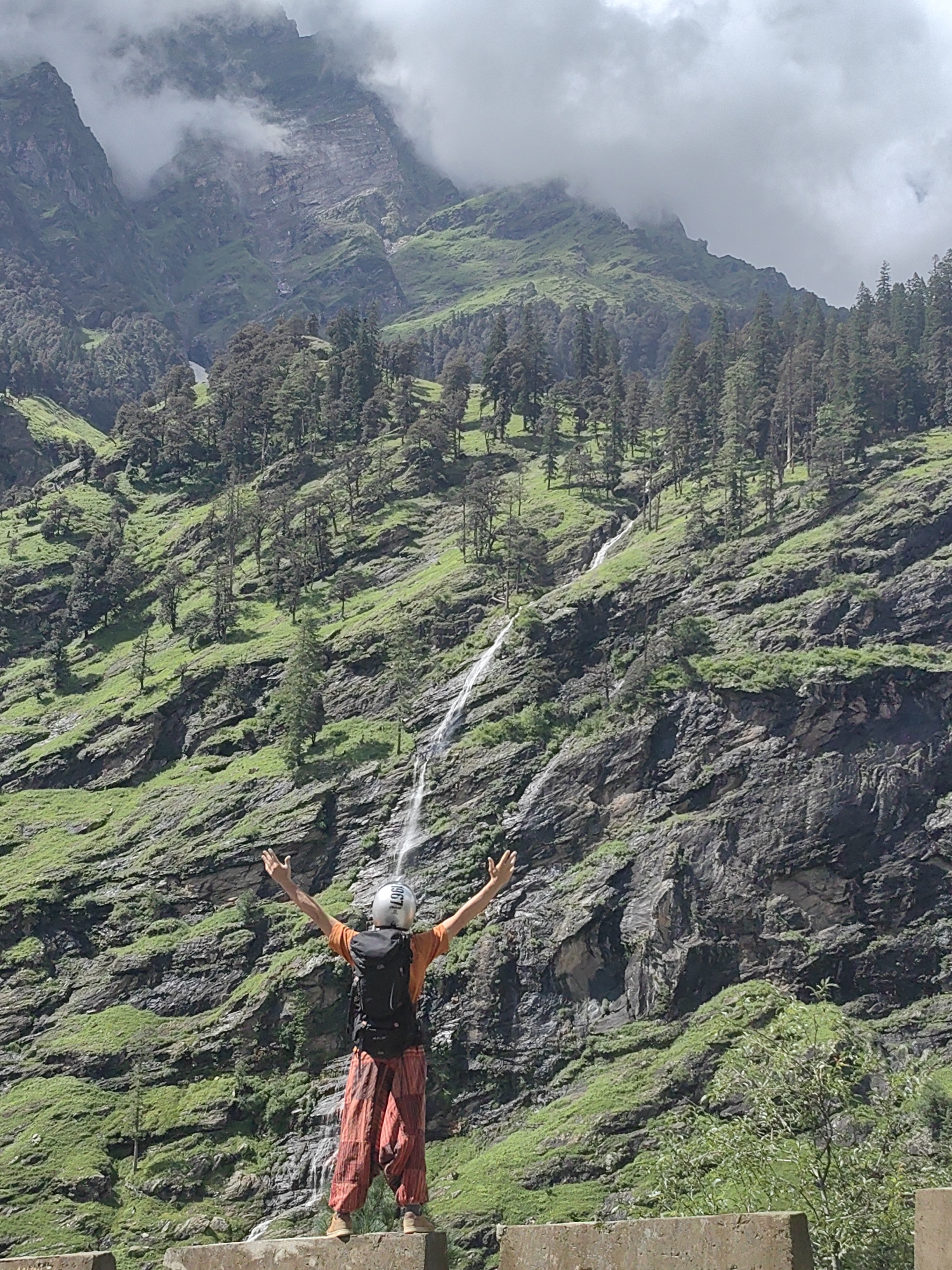 A person stands with arms outstretched on a stone barrier in front of a steep, lush green mountainside in Kothi, India. The person wears a silver helmet, an orange t-shirt, and loose-fitting, patterned orange pants. A black backpack is worn over the t-shirt. The mountainside is covered in vegetation and features a thin waterfall cascading down its rocky face. Dark green trees are scattered across the lower slopes, and the peaks of higher mountains are partially obscured by clouds. The sky is overcast with white and grey clouds.