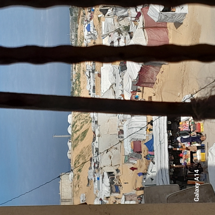 The image depicts an outdoor scene in the city of Khan Yunis, Palestinian Territory, viewed through three vertical, dark, irregular bars, likely from a window. The primary subject is an extensive, densely populated displacement camp established on sandy terrain under a partly cloudy sky.
In the foreground, a partial view of a light-colored wall or structure is visible on the bottom left, alongside a thin wire crossing vertically. The bottom center features a makeshift market stall with a white awning, displaying various goods such as packaged items and produce in crates. Several individuals, including adults and at least one child, are gathered around this stall, appearing to engage in shopping or observation. One person wears a white hoodie, another a dark top.
The midground is dominated by numerous temporary shelters and tents of varying sizes and materials, predominantly white or light-colored tarpaulins stretched over frames, with some darker, reddish-brown fabric structures. These shelters are densely packed, with sandy paths winding between them. Multiple individuals are visible traversing these paths. A person in a red garment walks on a path in the left midground.
In the background, a large sandy hill or embankment rises, exhibiting patches of green vegetation. Prominently situated on this hill are a large, circular, light-colored concrete structure (possibly a water tank or silo) on the left, and a tall, slender communication tower or utility pole to its right. Several smaller, white, dome-shaped structures are also present on the hilltop. The sky above is a mix of blue and darker grey cloud cover. Distant urban or natural features are faintly discernible on the horizon.