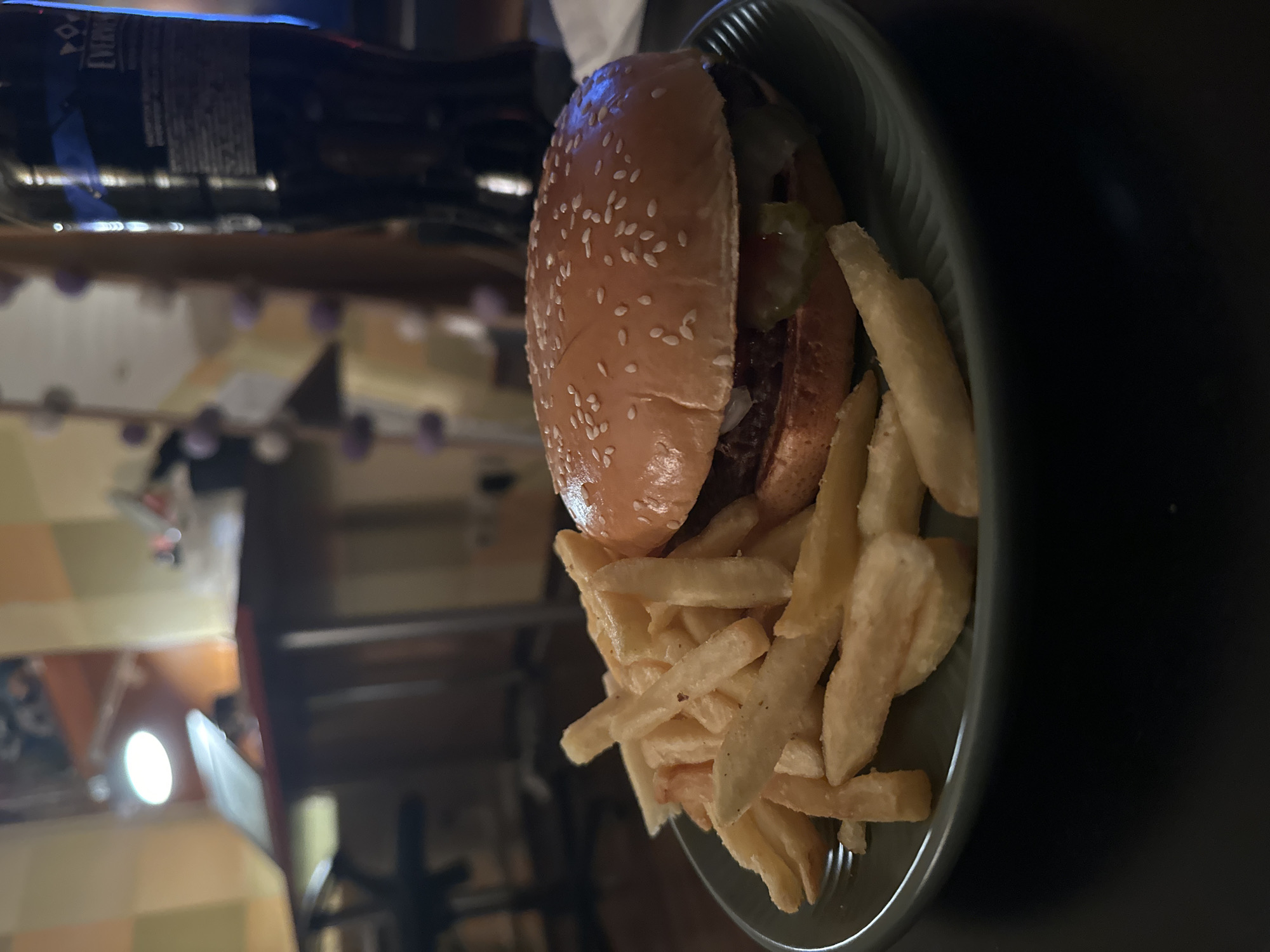 The image depicts a burger and fries on a plate, likely in a restaurant setting. A dark bottle, possibly a beverage, is visible behind the plate. The background is blurred, showing some indistinct furniture and lighting. There is no evidence of a location in Moscow, Russia.