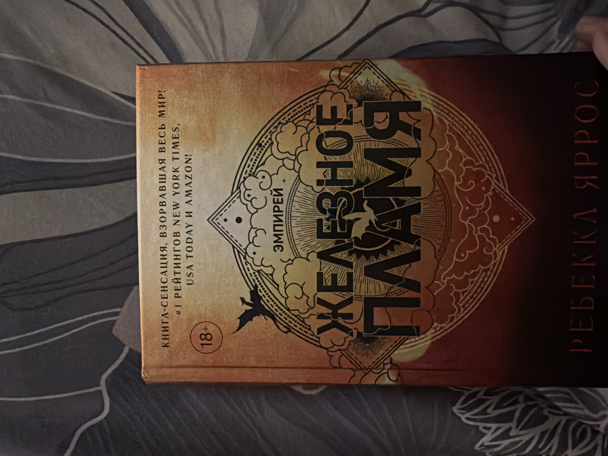 A book titled "Iron Flame" by Rebecca Yarros is lying on a bed. The text on the cover is in Russian. The cover is predominantly orange and brown. The bed has a grey and white patterned sheet. The image does not provide information about the location or city of Kazan, Russia.