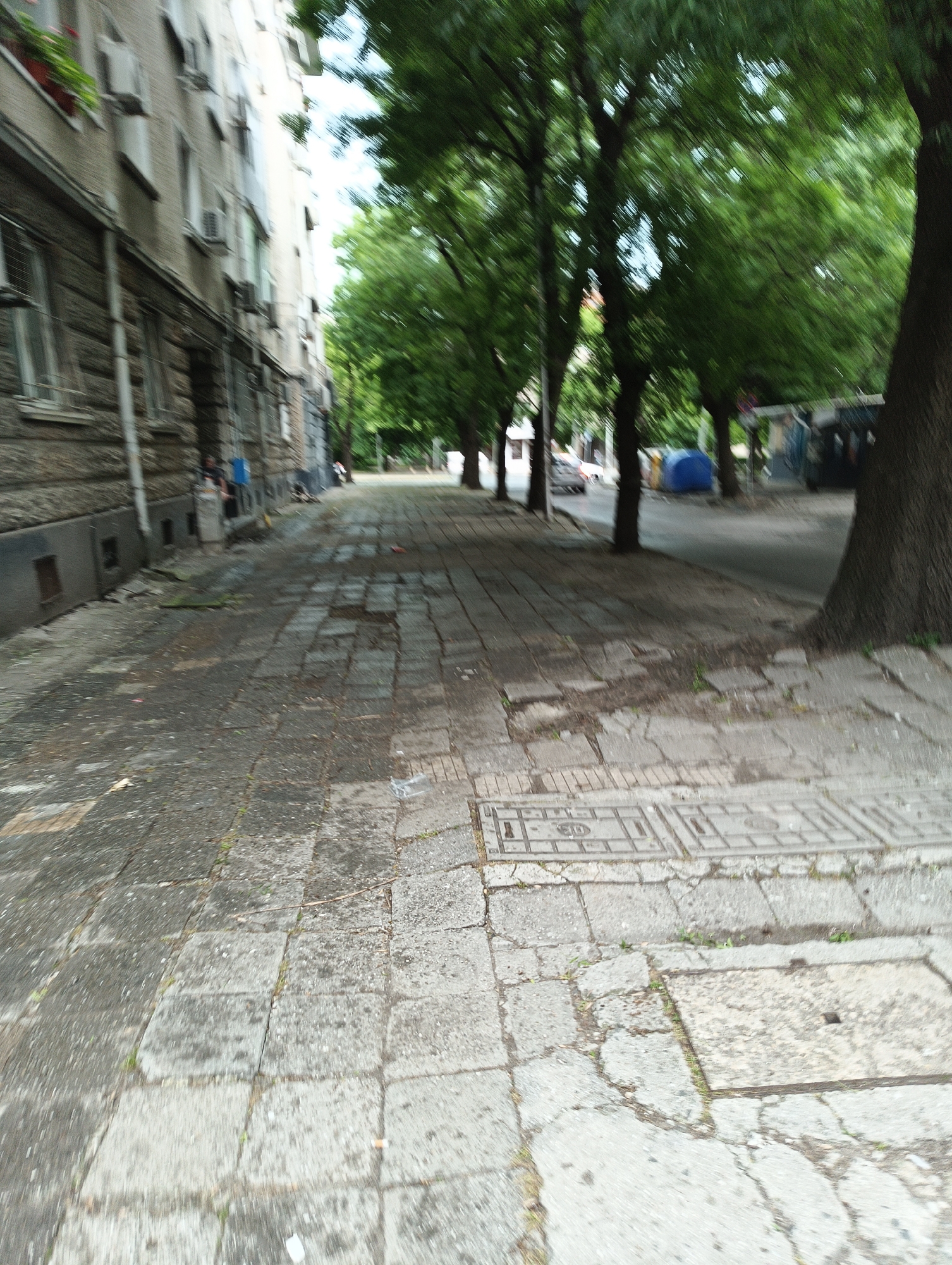 The image shows a street scene in Burgas, Bulgaria. The foreground is dominated by a gray, uneven sidewalk paved with concrete blocks, some cracked or displaced. A utility cover is visible. To the left, a tall, aged building with a stone facade is seen, with many windows and air conditioning units protruding. A person is sitting on the steps in front of the building. On the right, a row of trees lines the street, their foliage creating a green canopy. In the distance, the street continues, revealing other parked cars and temporary shelters or tents. The photo appears to be taken while moving, resulting in a slight blur, especially noticeable in the trees.