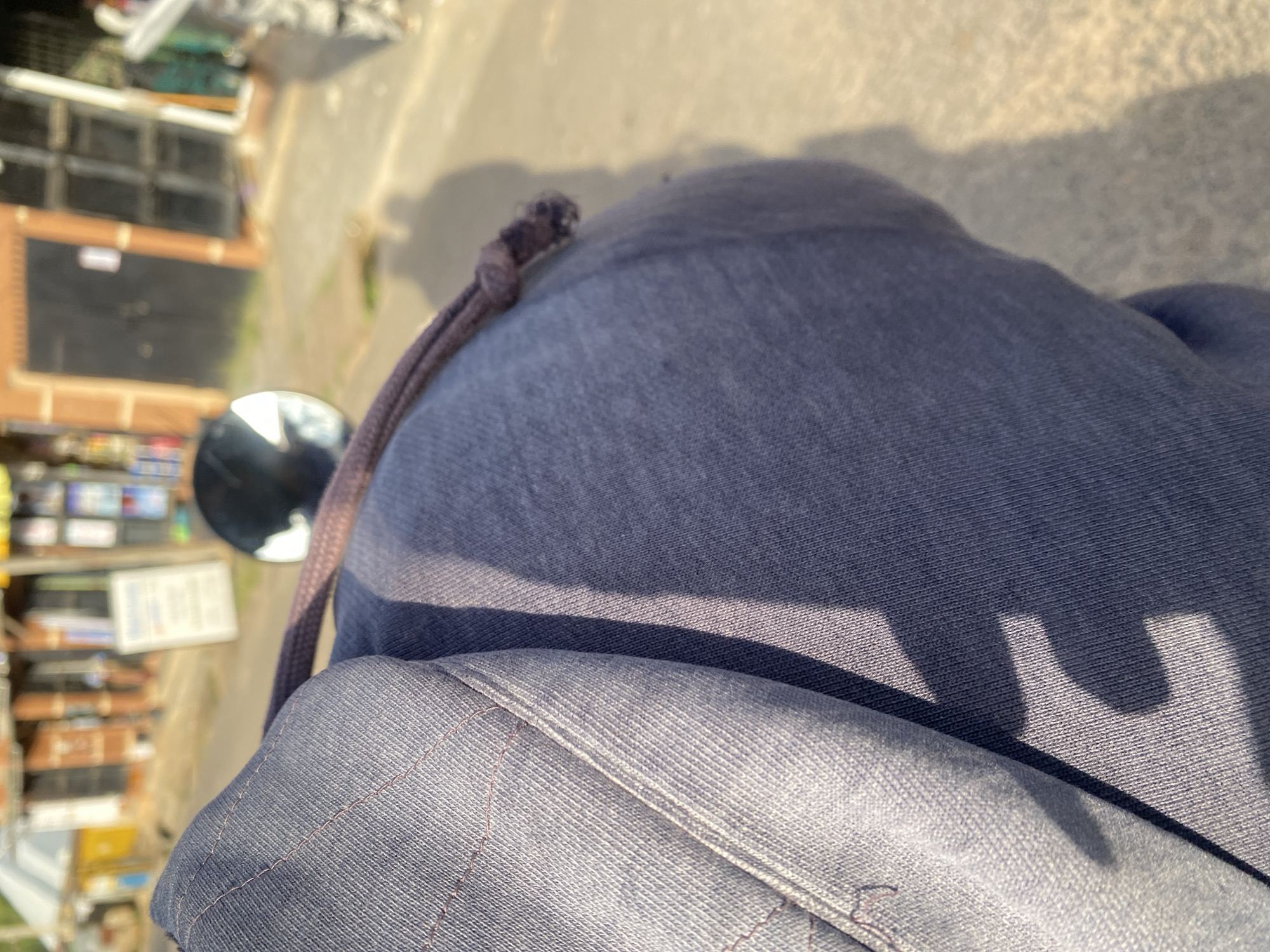 The image shows a close-up view of the shoulder and back of a person wearing a dark blue hooded sweatshirt. The sweatshirt has visible stitching and a drawstring with a knot. The background is out of focus but appears to be an outdoor scene, possibly a street in a city. In the distance, there are buildings with shop fronts and signage, consistent with a market or commercial area. The lighting suggests it is daytime, with sunlight casting shadows on the sweatshirt and the ground. Based on the context provided, this scene is located in the city of Jos, Nigeria.