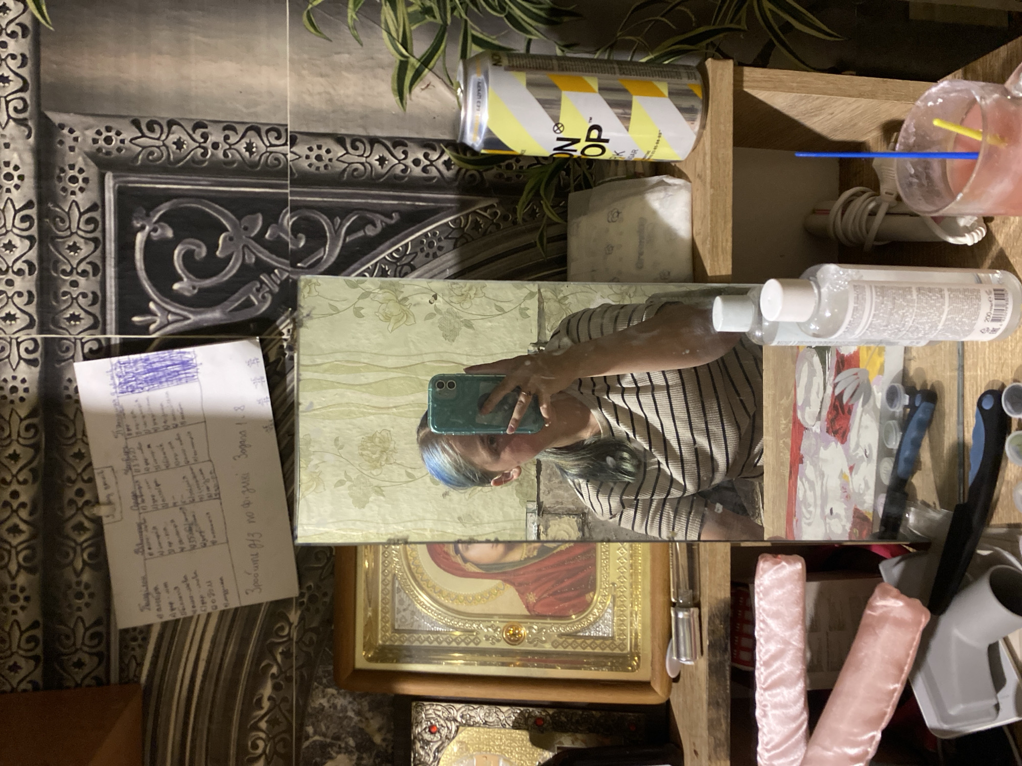A young woman with blue-tinted hair is taking a selfie in a mirror, holding a teal smartphone. She is wearing a striped t-shirt. The reflection shows her torso and head. In the background, several objects are visible on shelves. To the left of the mirror, an icon in a gold frame depicts a religious figure. Above it, a piece of paper with handwritten text is pinned to a patterned wall. To the right of the mirror, a can of "Lemon Drop" drink and a potted plant are on a shelf. Below the shelf, on another shelf, are two pink fabric-covered items, a grey plastic pipe fitting, and a blue-handled tool. In the foreground, on a lower shelf, is a clear mug with a pink liquid and two straws, as well as several small bottles. The overall scene appears to be an indoor setting, possibly a room or hallway, with a textured wall and wooden shelving units. The location of Nova Praha, Ukraine, is not indicated by any visual elements in the image.