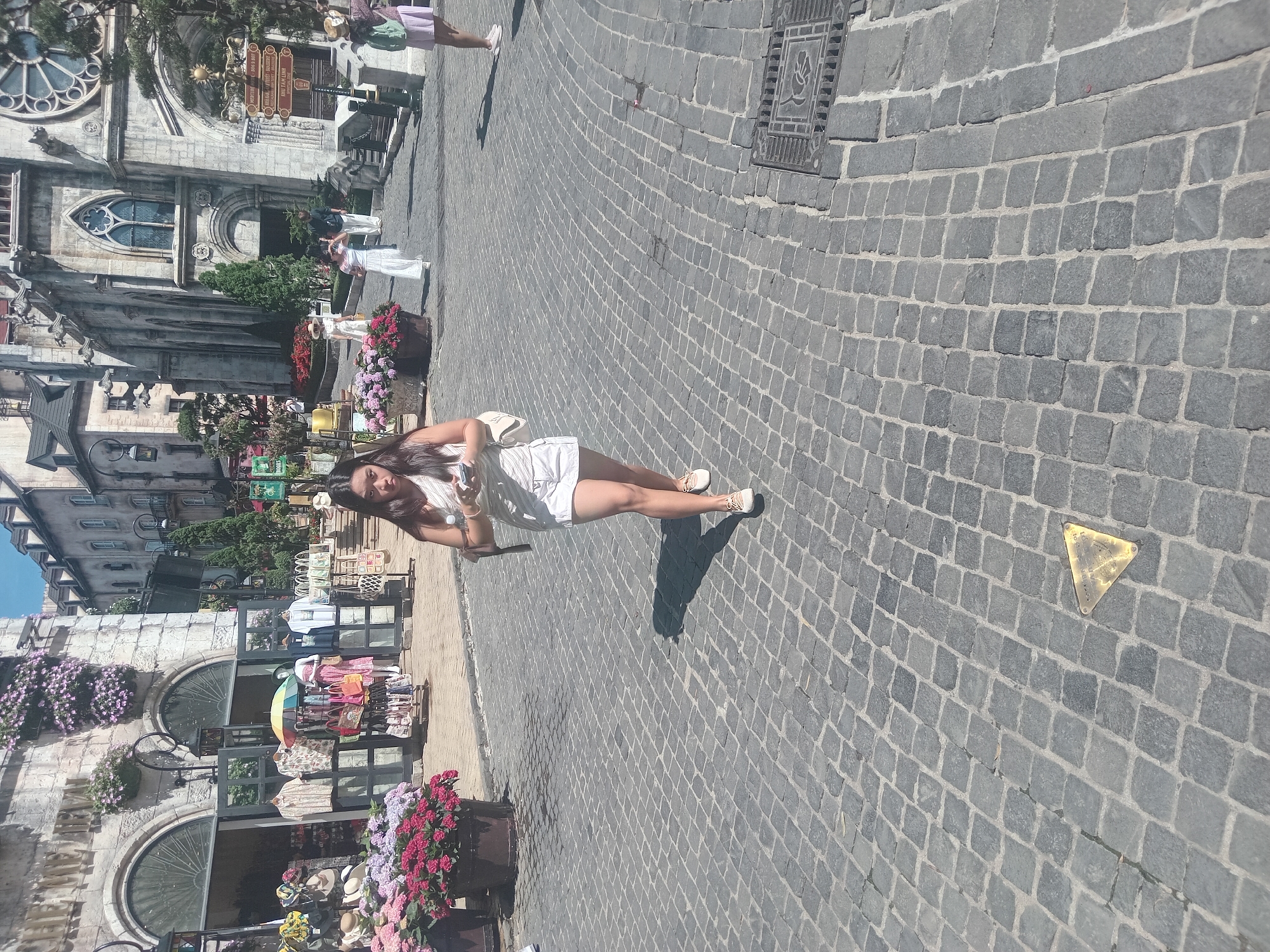 The image depicts a scene at Ba Na Hills, a tourist destination in Đà Nẵng, Vietnam, characterized by European-style architecture. In the foreground, a young woman is walking across a cobblestone street, holding a smartphone in her left hand and a small white purse in her right. She is wearing a light-colored sleeveless top and white shorts, with light-colored sneakers. The street is paved with dark gray cobblestones. In the midground, a shop with an open facade displays clothing and accessories, with potted flowers on the left side. In the background, a large stone building with Gothic architectural elements, resembling a church or castle, is visible. Other people are present in the background, and the sky is clear and blue, suggesting a sunny day. A triangular brass marker is embedded in the cobblestones in the lower right quadrant of the image.