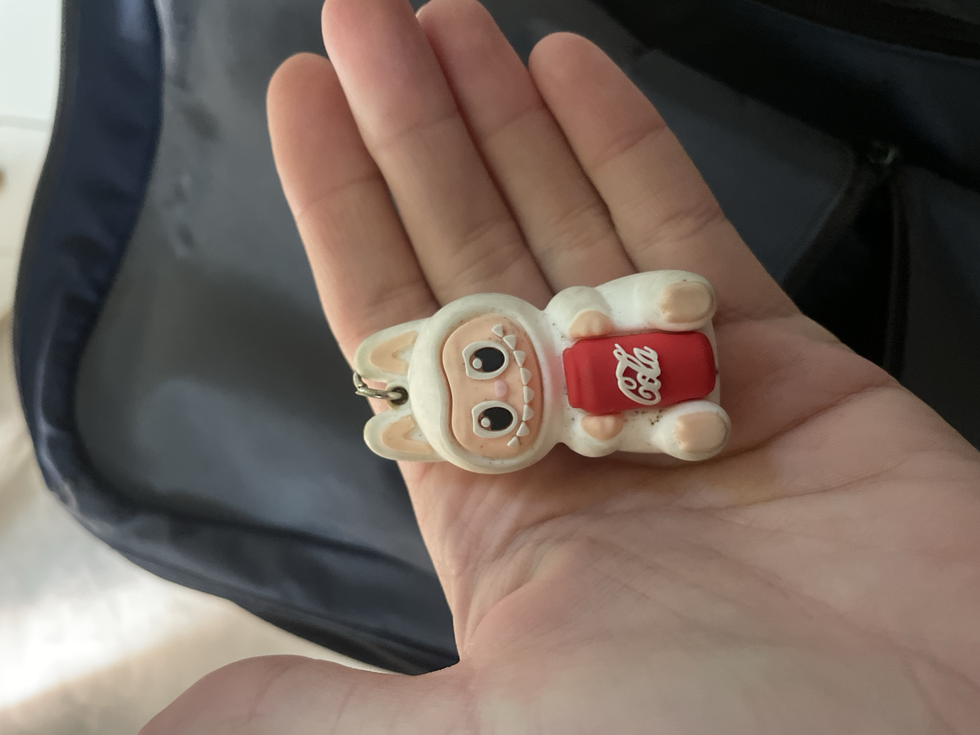 A close-up image shows a person's hand holding a white, anthropomorphic keychain figure. The figure features large, round black eyes, a small pink nose, and a wide-open mouth revealing several white, triangular teeth. It has slightly pointed ears and appears to be made of a soft, rubbery material. The character is holding a miniature red can with a distinctive white, stylized script that reads "Cola," strongly resembling the Coca-Cola brand logo. A small metal keychain ring is visible near the top of the figure's head.
The hand, with visible palm lines and skin texture, occupies a significant portion of the foreground. The background is a blurred dark blue or black fabric, likely part of a bag or backpack, showing subtle creases and reflections. A zipper element is faintly visible in the upper right background, suggesting the nature of the fabric item. The scene is brightly lit, emphasizing the details of the keychain and the hand. There are no specific cues within the image to indicate the time of day, weather, or the provided location context of Baranovka, Russia.