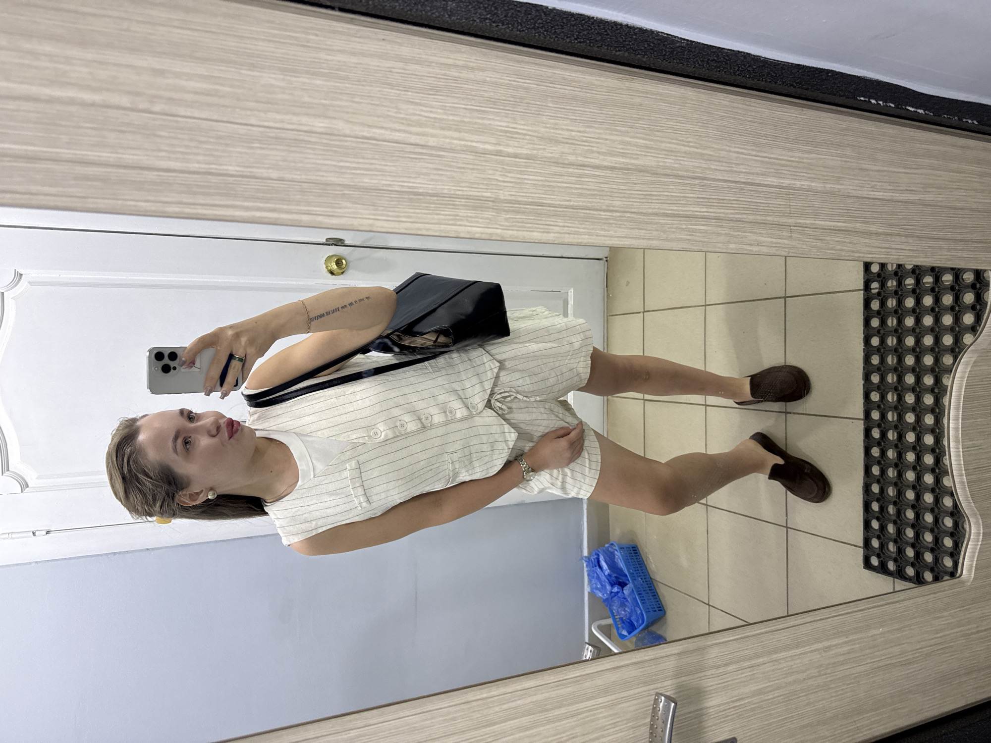 A woman is taking a selfie in a mirror. She is wearing a white striped romper, brown loafers, and carrying a black tote bag. The mirror is reflecting a bathroom with light-colored tile flooring and a blue plastic basket. The location is in the city of Chaikovsky, Russia. A black mat is visible in the reflection. The woman is holding a cell phone to take the picture. The wall visible in the mirror is a pale blue.
