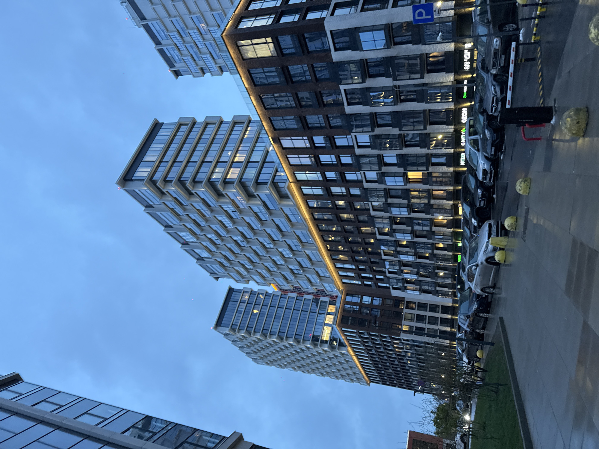 This image depicts a modern urban street scene in Khimki, Russia, captured during twilight, likely dusk. The setting features multiple high-rise buildings, a street with parked cars, and a clear, light blue sky.
In the foreground and extending along the right side of the frame, a multi-story building with a facade combining dark and lighter sections is prominent. This building is illuminated by warm yellow architectural lighting strips that accentuate its horizontal lines. Many windows display interior lights, indicating occupancy. On the ground level or reflected in its windows, commercial signage is visible, including "OZONE," "NOVO," "MVIDIO" (likely М.ВИДЕО, a large electronics retailer), and "APTEN A" (which appears to be АПТЕКА, Russian for pharmacy). Adjacent to this building is a paved area serving as a street or parking lot, where numerous cars, including sedans and SUVs, are parked parallel to the facade. A blue "P" parking sign, a black and white striped parking barrier, and yellow spherical bollards mark the area.
To the left and slightly behind, another tall, contemporary building stands out with its predominantly glass and lighter grey/white facade. This structure features a distinct angular and geometric design, creating a textured or stepped appearance. A third, similarly modern building is partially visible in the bottom-left corner.
The sky is a serene, light blue, suggesting the transition hour between day and night, without any visible clouds. The overall scene conveys a sense of modern urban development and architecture. There are no people visible, and no specific activities are taking place, presenting a quiet moment in a contemporary city environment.