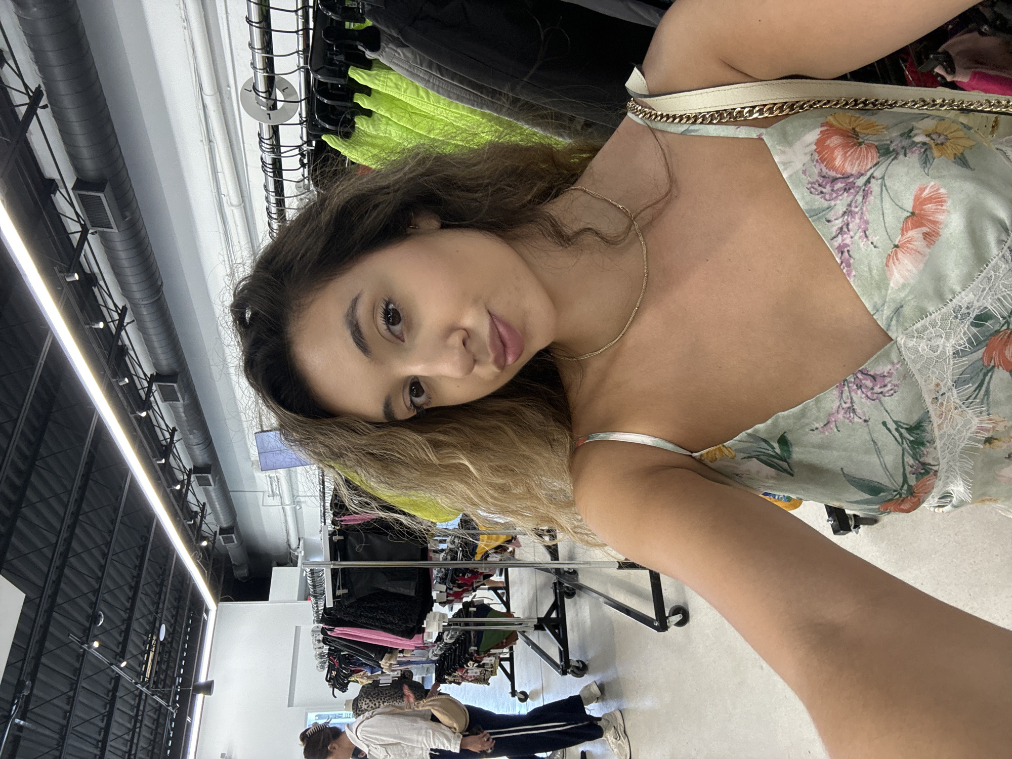 A young woman is taking a selfie in what appears to be a clothing store in a brightly lit commercial building. The woman has brown hair, tan skin, and is wearing a floral patterned top with a gold necklace. Behind her are clothing racks filled with various articles of clothing. Other people are visible in the background, further back in the store. The location is indicated to be in Miami, United States of America. The building has an industrial ceiling with exposed pipes and lighting fixtures.