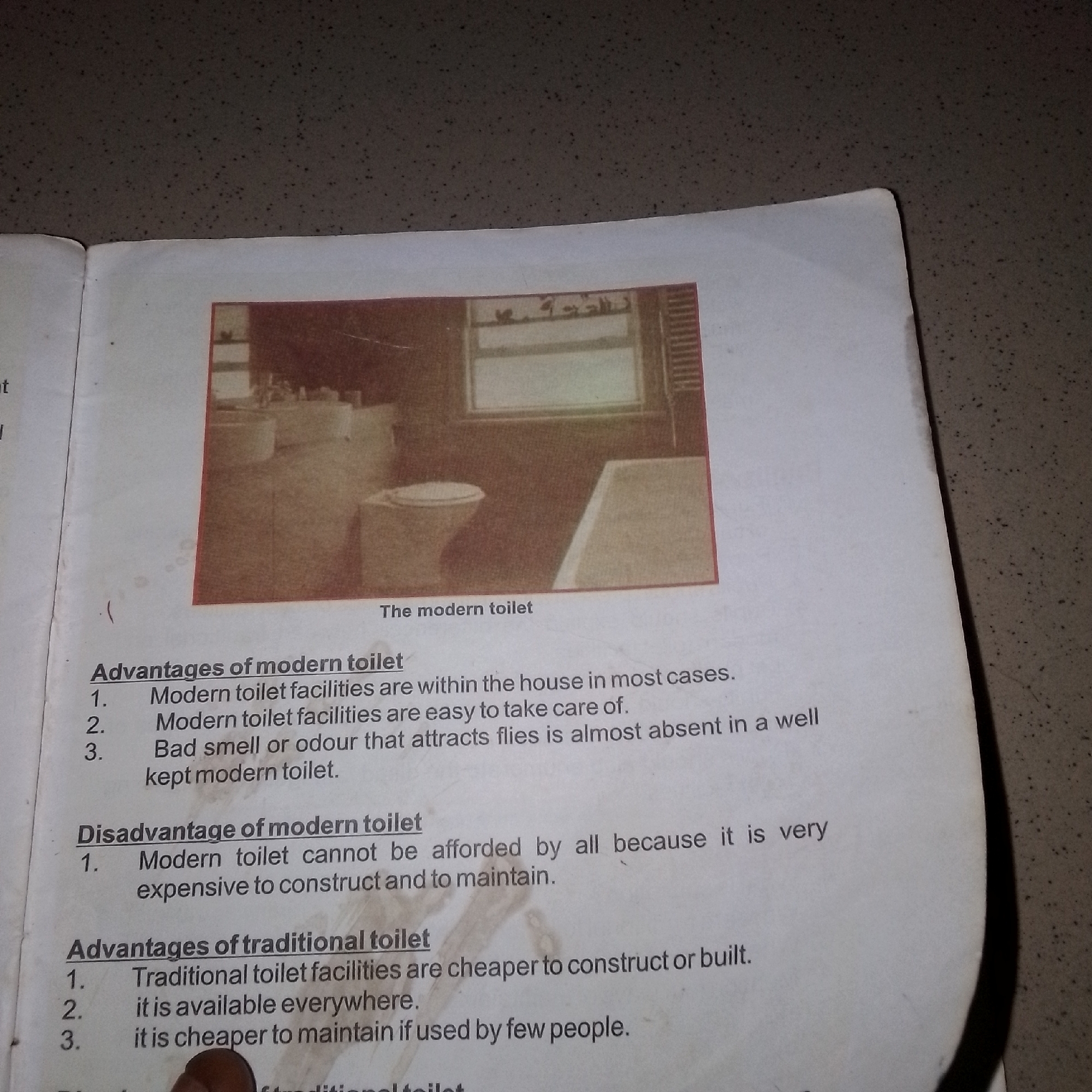 The image shows a page from a book with text and a photograph of a modern toilet. The text discusses the advantages and disadvantages of modern and traditional toilets. The photograph depicts a clean, tiled bathroom with a toilet and a sink. The image is taken from an angle that shows the top of the toilet and the sink. There are no people in the image. The background appears to be a wall with a window. The book page itself is white and slightly yellowed, suggesting it is an older publication. The surrounding environment is a textured, speckled surface that appears to be a table or desk. There is no information in the image that links it to Jalingo, Nigeria.