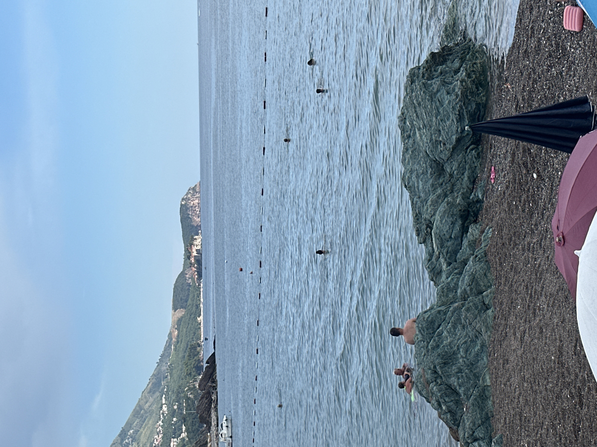 This image captures a serene scene at a beach in Rafailovići, Montenegro. The sky is a clear, bright blue, suggesting a sunny day, likely in the late morning or early afternoon. The sea is calm with gentle ripples, and a safety line with buoys stretches across it, delineating a swimming area.
In the water, several people are visible, some swimming and others floating. On the rocky shore to the right, two men are sitting on the rocks, with one appearing to be in conversation with the other. Further to the right, on the dark pebble beach, two umbrellas are set up, one black and one reddish-purple, offering shade. There are also hints of beach towels and bags.
The backdrop features a lush, green mountainside dotted with white buildings, characteristic of coastal Mediterranean towns. The overall mood is one of relaxation and leisure, as people enjoy the pleasant beach weather. No visible text is present in the image.