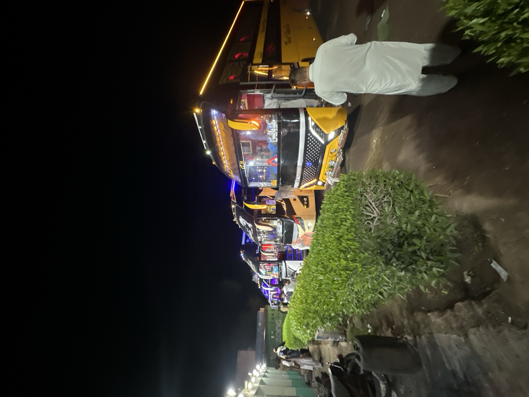 At night in an urban location in Unknown, Unknown, a row of parked buses is visible. They are illuminated with various lights. One individual in a white outfit is near one of the buses. Vegetation is interspersed among the buses. Further down the row, other people are also present, and a few motorcycles are parked nearby. The sky is dark.