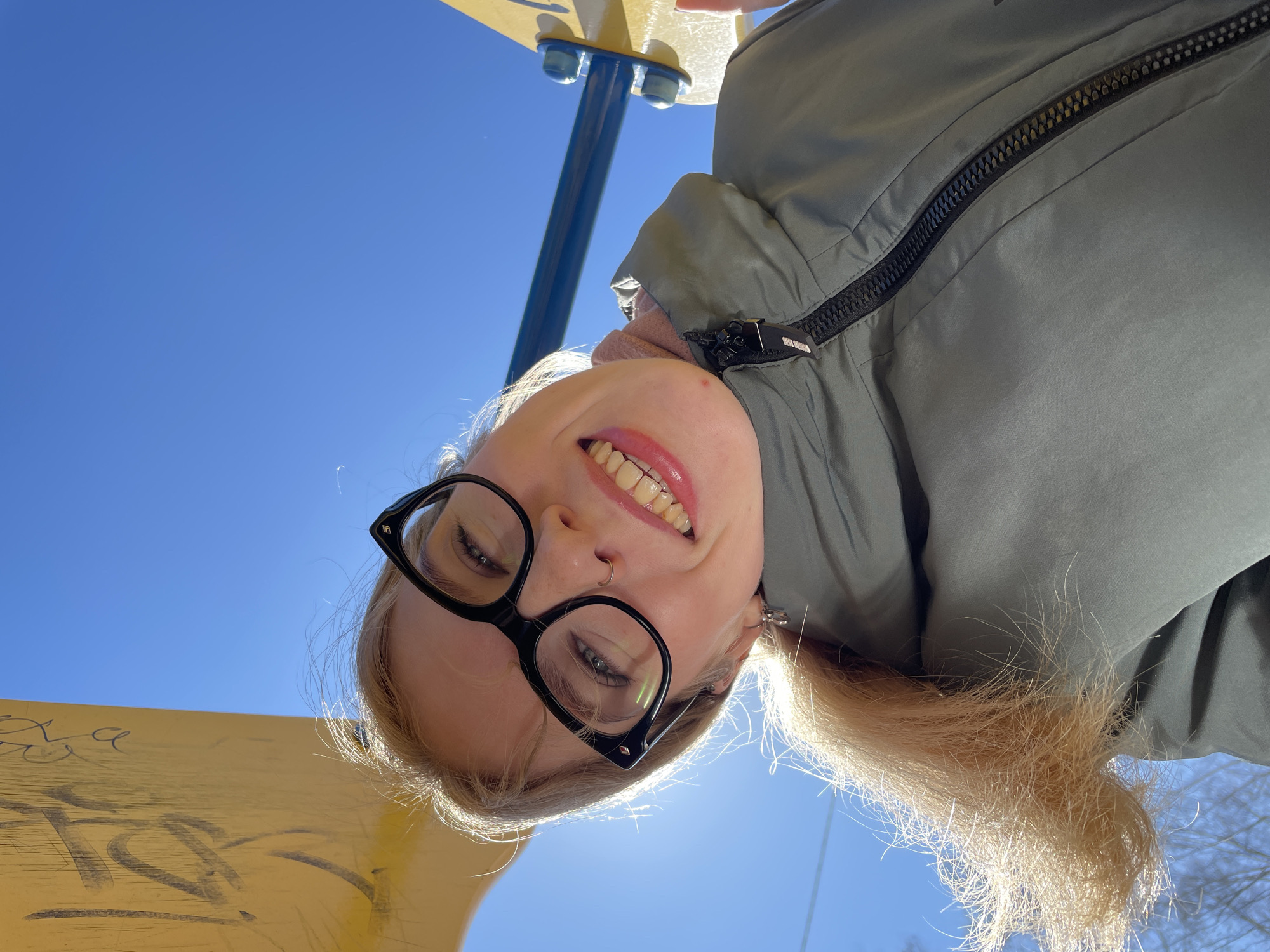 A fair-skinned woman with blonde hair and black-framed glasses is positioned in the foreground, looking upwards. Her face is illuminated by sunlight. She wears a green jacket with a visible zipper. A piercing is noticeable in her nose. Behind her, the clear blue sky fills much of the background. Elements of a yellow playground structure are visible on the left. Trees are faintly seen in the far background. The location is Izhevsk, Russia.
