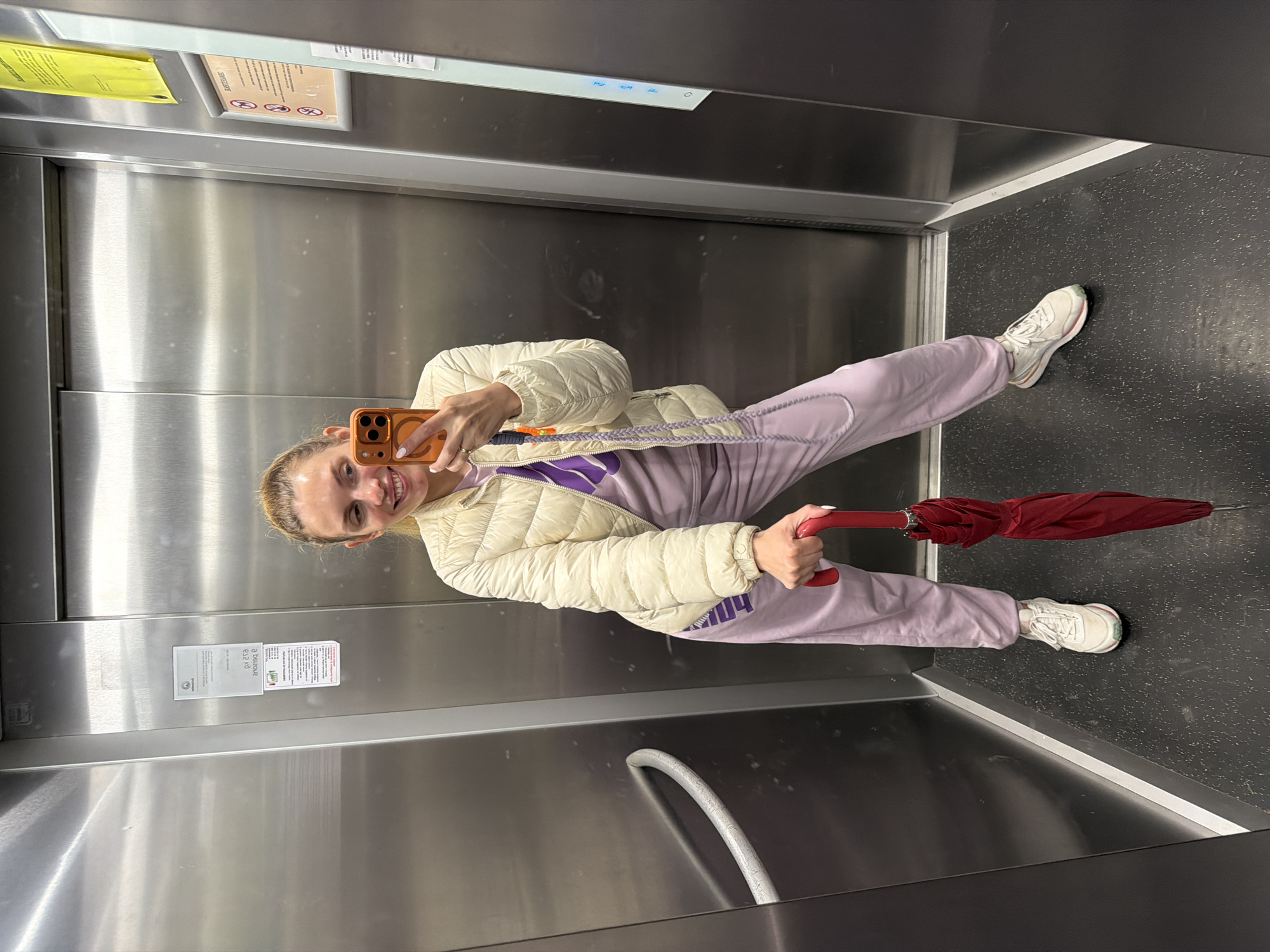A woman is taking a selfie in an elevator. She is wearing a white puffer jacket, light purple sweatpants, and white sneakers. She is holding a red umbrella in her left hand and a smartphone in her right. The elevator has stainless steel walls and a dark gray floor. There are informational signs posted on the elevator walls. While the image itself does not explicitly confirm the location, it is possible to infer the city of Budva, Montenegro based on metadata not included in the prompt.