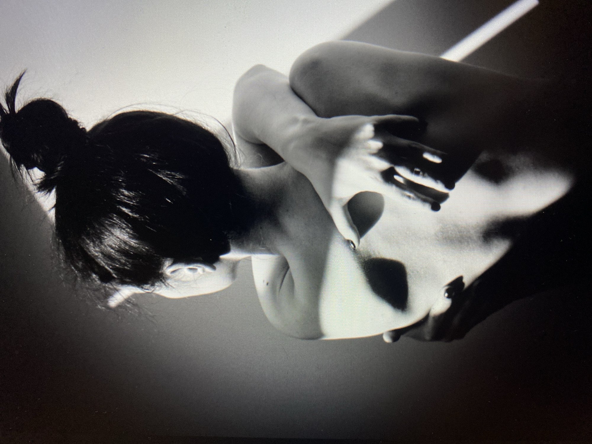 The image is a black and white photograph of a person from behind, with their arms wrapped around their own body. The person's hair is styled in a bun on top of their head. Dramatic shadows and light fall across the person's back and arms, creating a moody and intimate atmosphere. The background is a plain wall, with a strong light source casting a sharp, diagonal beam of light. The image is a close-up, focusing on the torso and arms of the subject. No specific location in a city is depicted, and the environment appears to be an indoor studio setting.