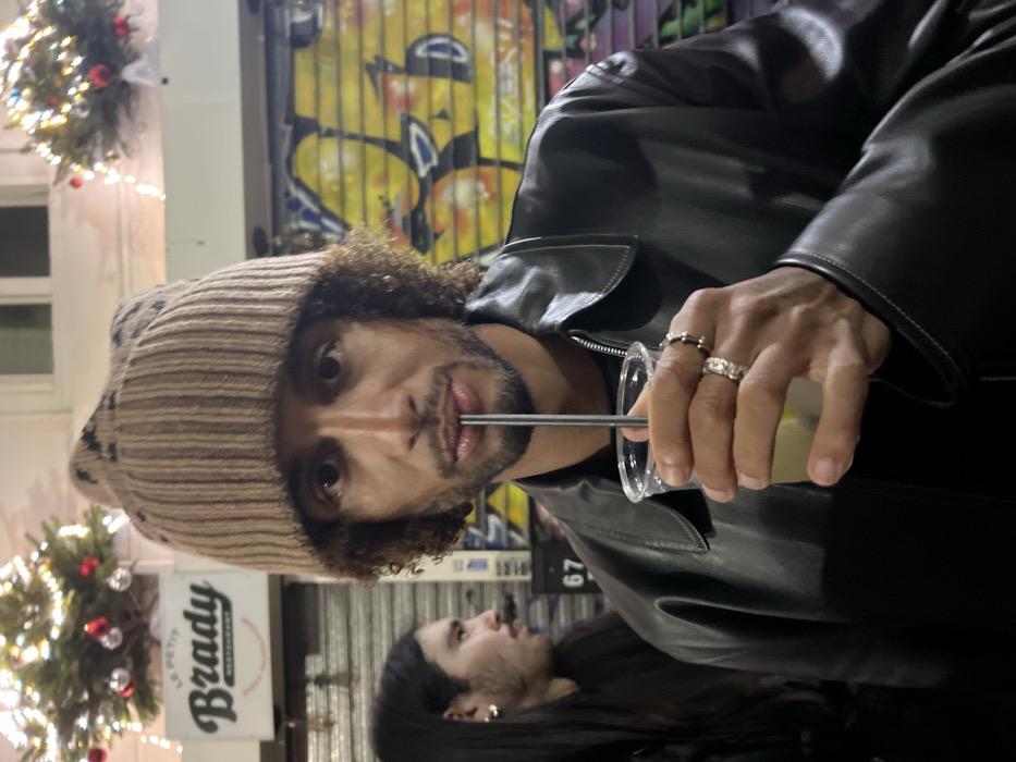 A man wearing a brown knit hat and a black leather jacket drinks from a clear plastic cup using a metal straw. He has curly hair and wears rings on his fingers. Behind him are street lights, a sign that reads "Brady Restaurant," and another person with long dark hair. A graffiti-covered storefront provides a colorful backdrop.