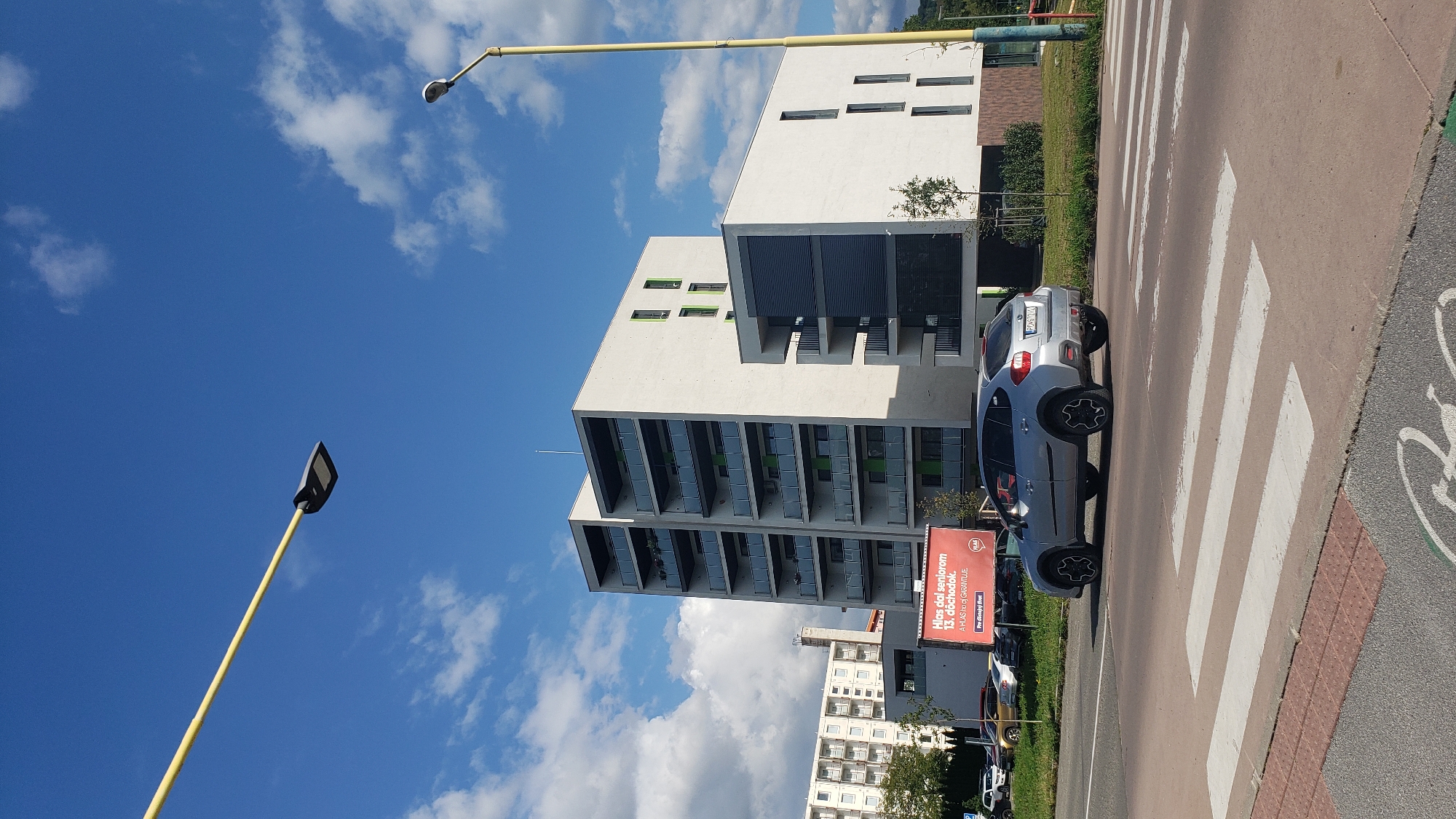 A silver Subaru XV is crossing a pedestrian crossing on a street in Prešov, Slovakia, on a sunny day with a partially cloudy blue sky. The car is moving from left to right in the frame, with its rear facing the camera. The pedestrian crossing is marked with white stripes and textured red and grey paving.
In the background, a modern, multi-story apartment building with white facades, green accents, and balconies with glass railings dominates the scene. To the left of this building, another apartment building, appearing older and with a uniform facade of white windows, is partially visible. A red billboard with text in Slovak, which appears to be an advertisement for a political candidate named "HLAS", is situated in front of the apartment buildings.
Two streetlights are visible in the image. One is a modern, dark-colored lamp on a yellow pole extending from the upper left corner of the frame. The second is a more traditional lamp on a yellow pole on the right side of the image. Vegetation, including trees and shrubs, is present along the side of the road and in front of the apartment buildings. Other vehicles and a parking lot are visible behind the billboard. The overall impression is of an urban environment with modern residential architecture and public infrastructure.