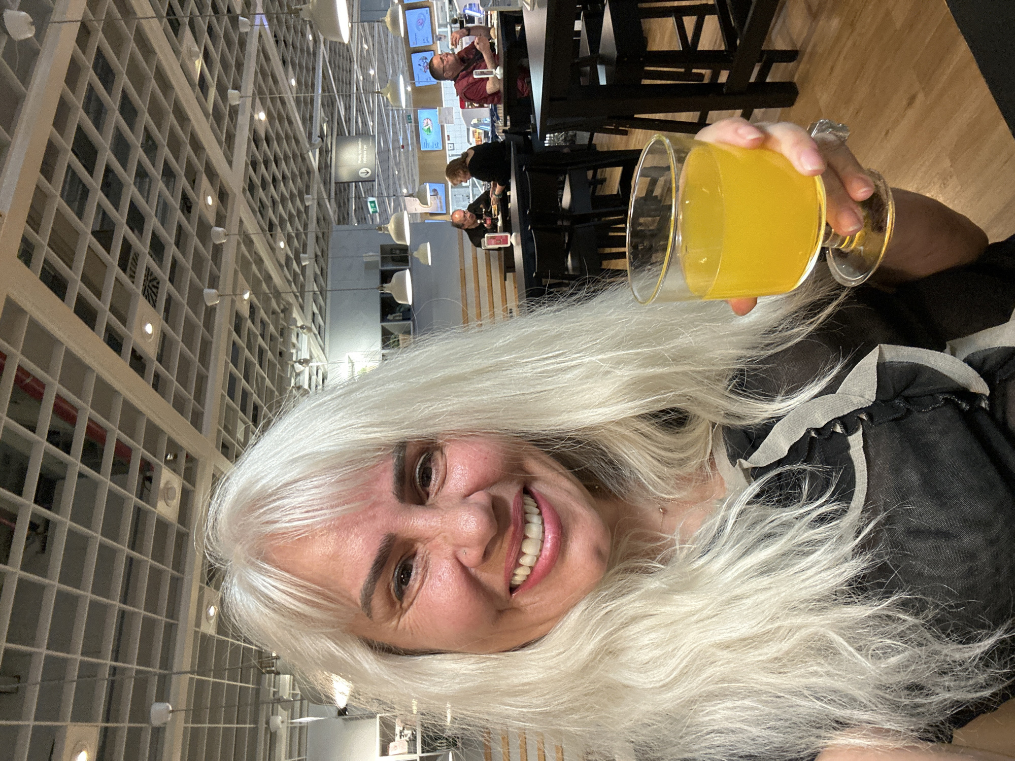 A woman with long, white hair is holding a glass of yellow liquid, smiling at the camera. She is wearing a dark-colored top. The background features a restaurant setting, with tables and people seated in the distance. The ceiling has a distinctive grid pattern with lights. A sign is visible in the background. The location appears to be within a large commercial space, possibly a furniture store with a dining area. It is not possible to identify the precise location within Sofia, Bulgaria without additional information or landmarks.