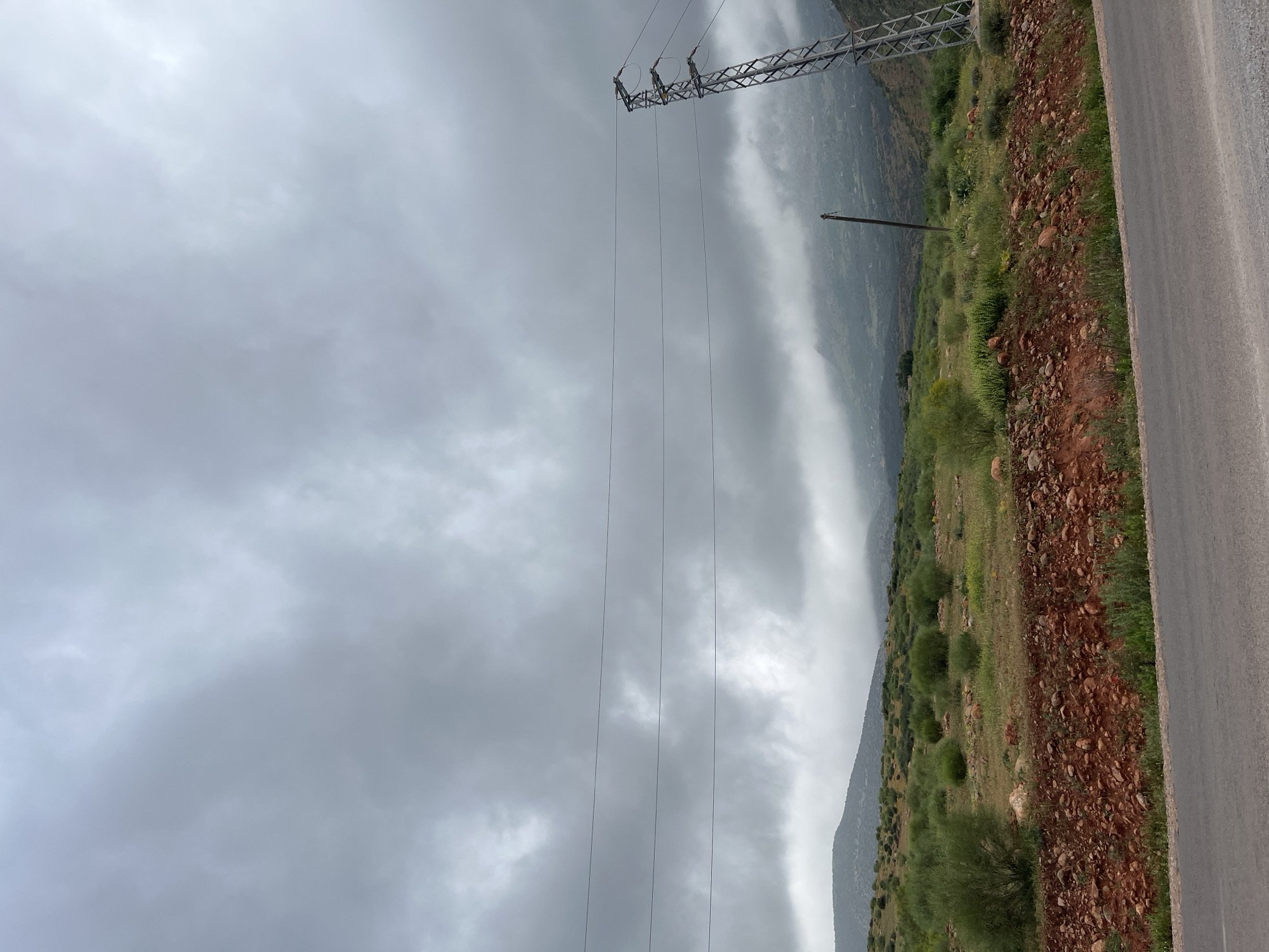 The image depicts a roadside view in Ouaouizert, Morocco, characterized by overcast weather and a mountain landscape. The sky is filled with grey clouds. A utility tower with power lines runs diagonally across the frame. A mountainside covered in vegetation, consisting of grass, bushes, and reddish-brown earth with rocks, slopes down to the left. A grey asphalt road borders the vegetation on the right. Another lone utility pole rises next to the mountainside.