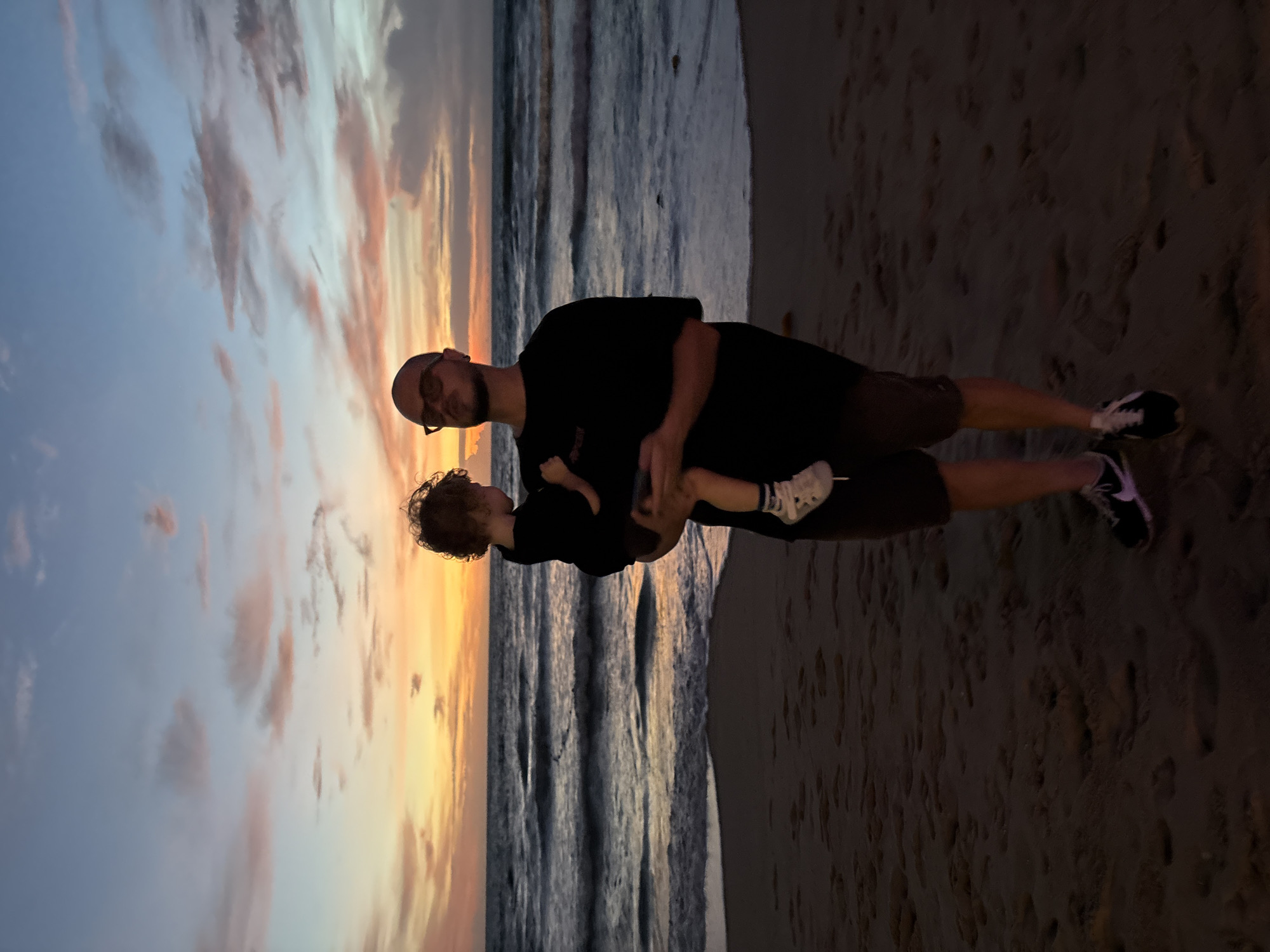 At dusk on a sandy beach in Choeng Thale, Thailand, a man is holding a young child. The man, who is wearing glasses and a black t-shirt, is facing forward while looking at the child. The child, facing away from the camera, is being held in the man's arms. The ocean is in the background with waves breaking on the shore. The sky is filled with clouds in shades of orange, pink, and blue, indicating the sunset. The sand on the beach shows footprints.