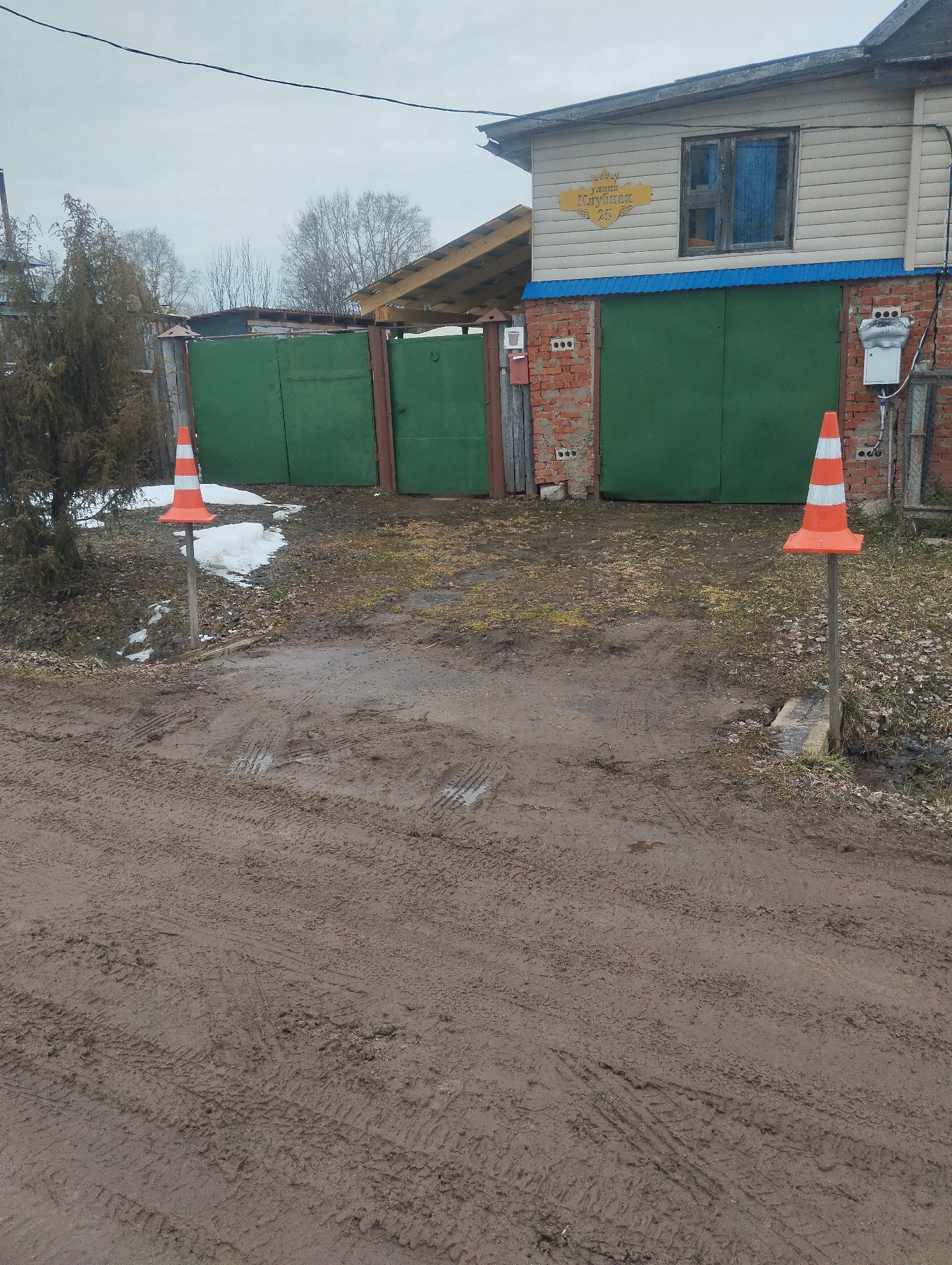 The image shows a muddy driveway in front of a green gate and a two-story building. The address on the building reads "Klubnaya, 25," indicating the location is in Russia, likely Grishanki. Two orange and white traffic cones are placed on posts on either side of the driveway entrance. Patches of snow remain near the left cone and along the base of the fence. Tire tracks are visible in the mud. The sky is overcast. The building is constructed of brick on the lower level and siding on the upper level. A partially-obscured wooden shed or structure is visible behind the green gate on the left side. A small coniferous tree stands to the left of the entrance.