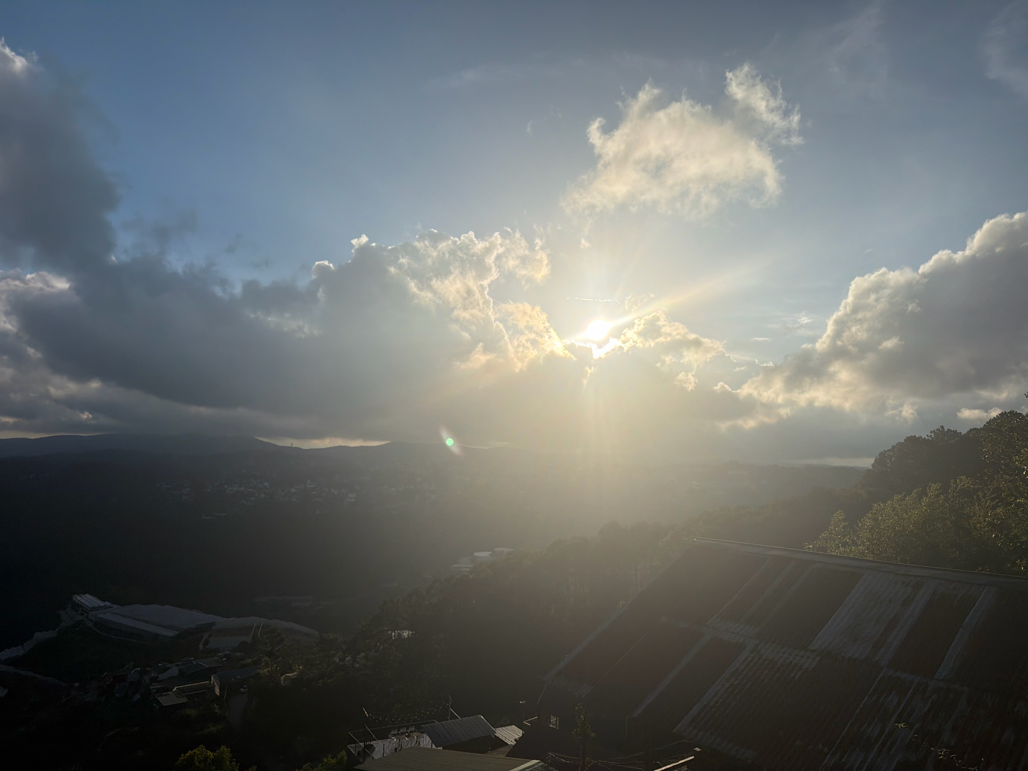 The image depicts a sunlit landscape with a view of a valley and distant hills under a partly cloudy sky. The sun is positioned high in the sky, breaking through a cluster of clouds and casting bright rays that create a lens flare effect. In the foreground, the corrugated metal roof of a building is visible, angled downwards and showing signs of age. The middle ground features a lush green valley with scattered buildings and vegetation, including what appears to be a large industrial or agricultural structure with a white roof on the left. The background consists of rolling hills obscured by a haze and the dimming light of the sun. The scene suggests a rural or semi-rural setting with a sense of elevation, possibly from a hillside. The time of day appears to be late afternoon or early morning, given the angle of the sun and the quality of light. The overall atmosphere is serene and natural, with a touch of human habitation.
