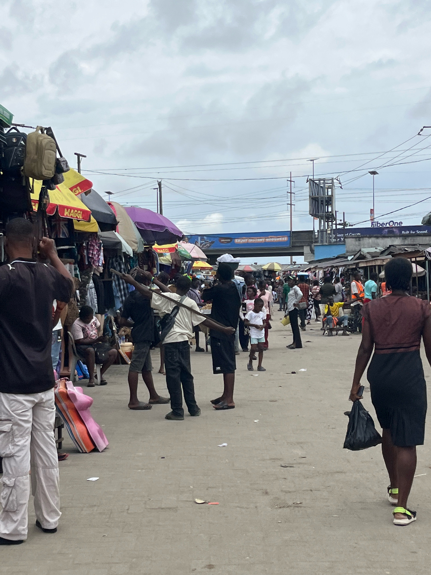 The image depicts a bustling outdoor market scene in Ado, Nigeria, under an overcast sky. In the foreground, a person wearing a black t-shirt and light-colored cargo pants is seen from the waist down, with their back to the camera. To their right, a woman in a dark brown top and black skirt walks away from the camera, carrying a black plastic bag in her right hand and a black smartphone in her left. She is wearing neon yellow and black sandals.
Further into the market, several individuals are engaged in various activities. Some are standing and interacting, while others appear to be browsing or selling goods. A group of men are visible, some holding sticks or poles. Stalls are set up with clothing and other merchandise displayed under colorful awnings.
The environment is a dusty, open area, likely a street or market square, with concrete paving. Overhead, power lines and poles crisscross the sky. In the background, a large structure with scaffolding and signage, including a "FiberOne" advertisement, suggests the presence of infrastructure. The overall impression is one of a vibrant and active marketplace in an urban Nigerian setting.