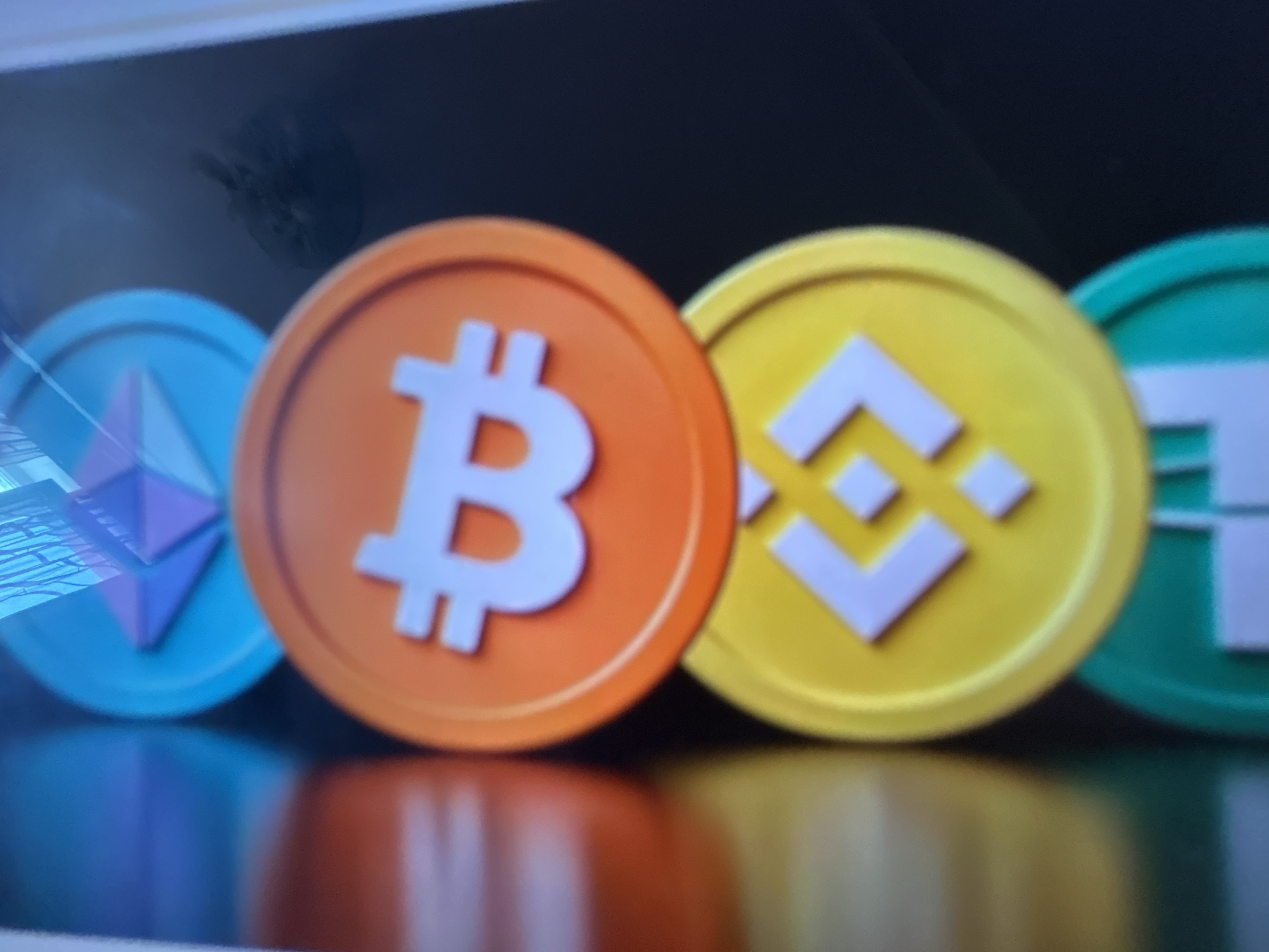 The image displays a close-up, slightly blurred shot of several cryptocurrency coins lined up against a dark background. From left to right, the coins visible are Ethereum, Bitcoin, Binance Coin, and Tether. The Ethereum coin is blue with a purple diamond-shaped logo. The Bitcoin coin is orange with the white Bitcoin symbol. The Binance Coin is yellow with a white Binance logo. The Tether coin is teal with a white "T" symbol. The coins are reflective, casting a distorted reflection on the dark, glossy surface they are resting on. The overall scene appears to be a digital rendering or a still from a video, possibly intended for promotional or informational purposes related to cryptocurrencies. There is no discernible activity or event taking place, nor are there any specific location cues, time of day, or weather indicators. The focus is solely on the distinct visual representation of these popular cryptocurrencies.