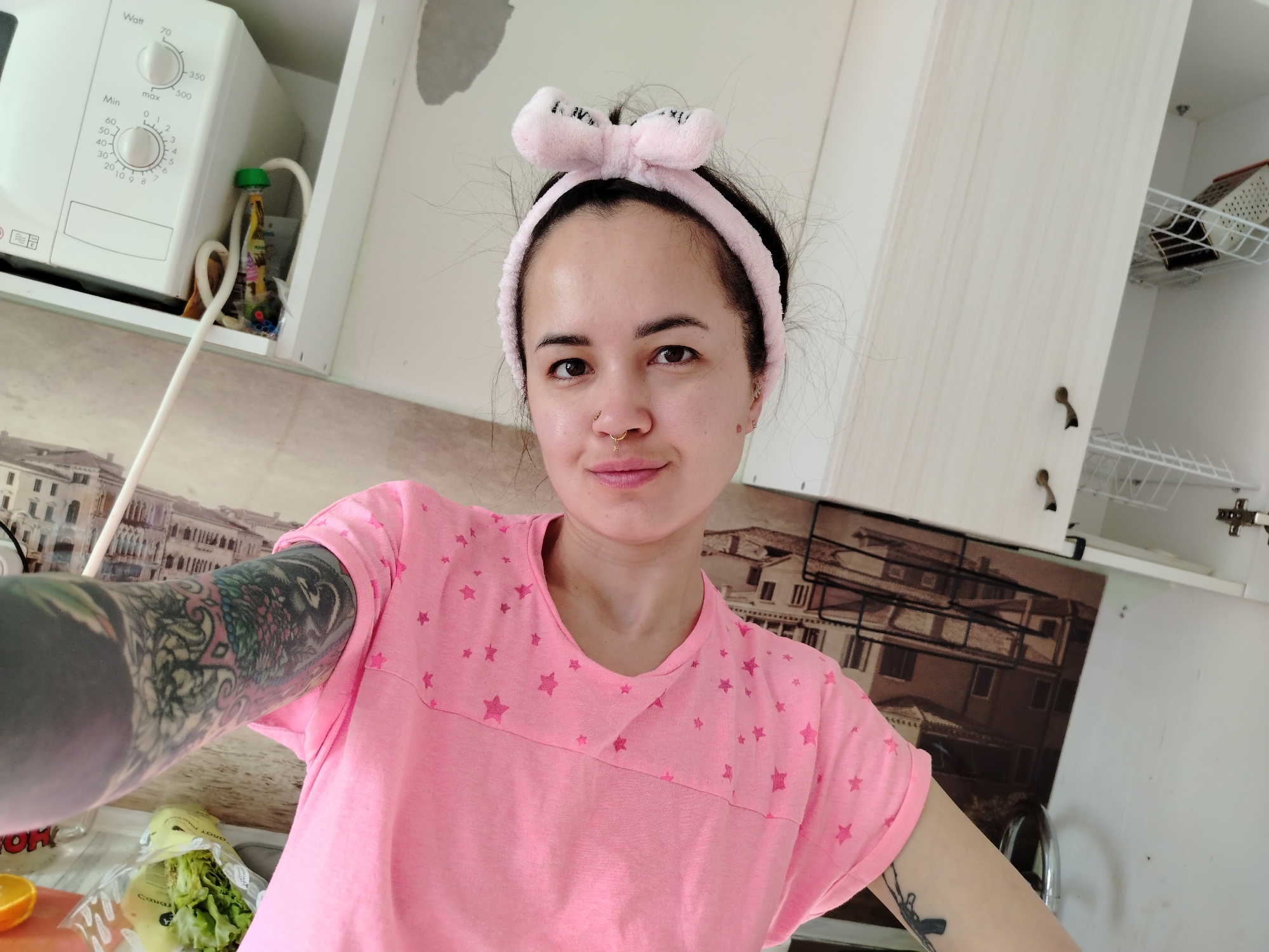 A person, likely female, takes a selfie in a kitchen. The person has dark hair, fair skin, and is wearing a pink headband with a bow. They are also wearing a pink t-shirt with star patterns. The person has several tattoos on their arms.
The kitchen features white cabinets and a backsplash with an image of a cityscape. A white microwave is visible on the upper left. Various food items, including an orange and a package of lettuce, are on the counter. The setting appears to be in a home.
The cityscape on the backsplash appears to be a photograph of Venice, Italy. The location in the city of Noviy, Russia is not identifiable from this image.