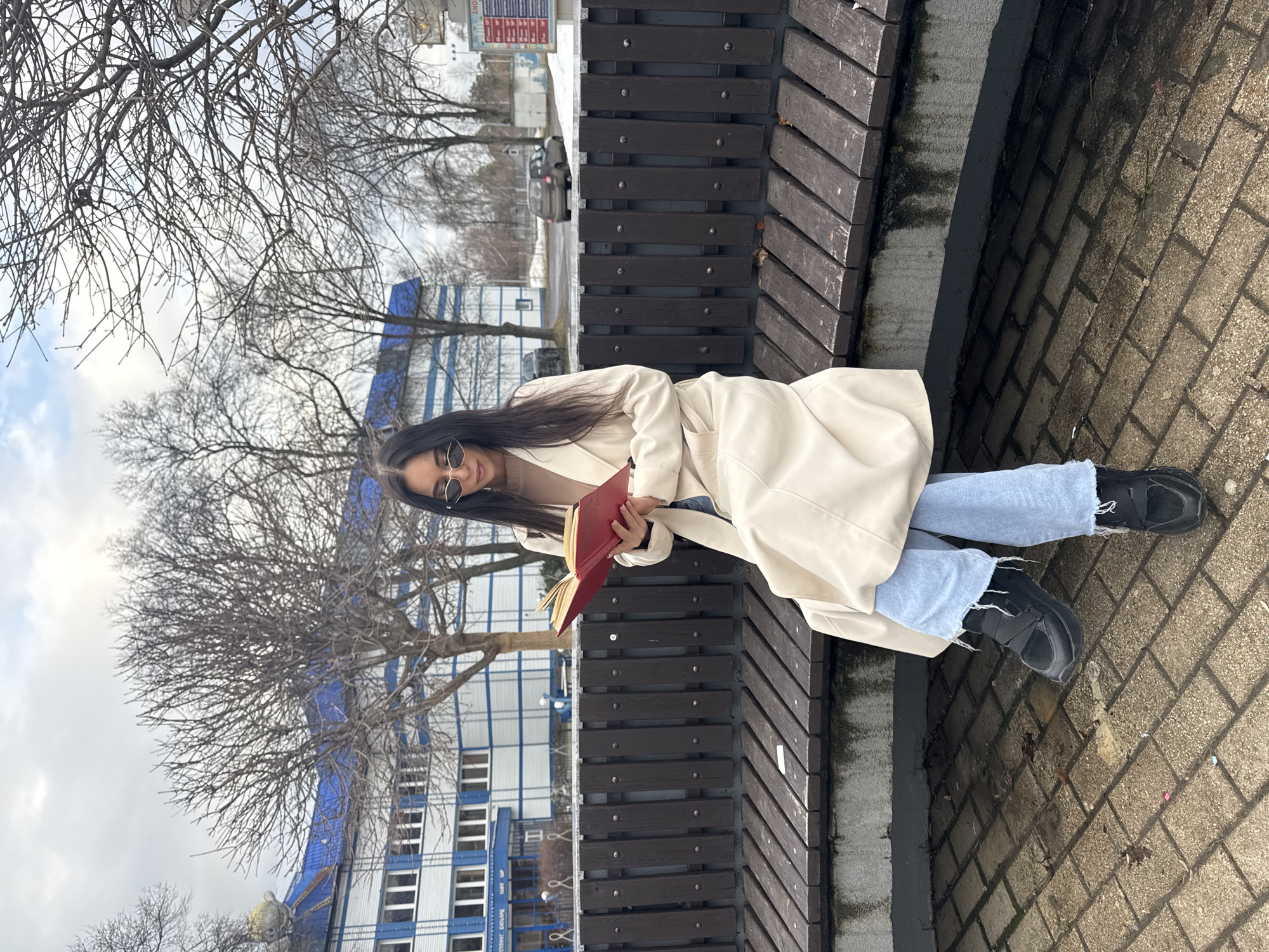A woman with long dark hair and sunglasses sits on a wooden bench, reading a book. She is wearing a light-colored coat and light blue jeans with black shoes. The bench is situated on a paved surface, possibly a sidewalk. In the background, there is a building with blue and white accents and bare trees. A street with some parked cars is also visible in the background. The image depicts a scene in the city of Chaikovsky, Russia.