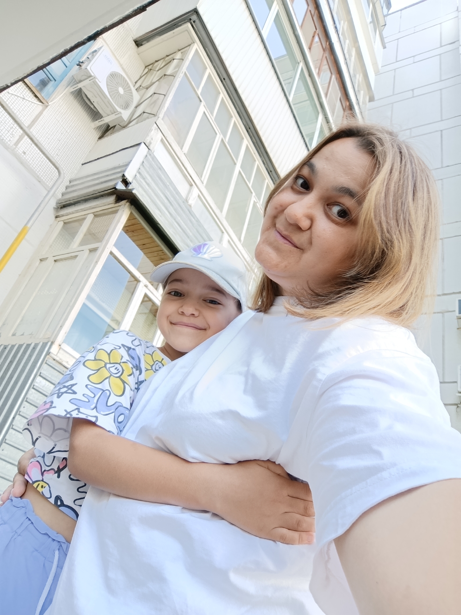 A medium shot shows a woman and a child from a low angle outside a building in Chaikovsky, Russia. The woman is in the foreground, holding the child. Both are smiling at the camera. The child is wearing a light blue cap and a white shirt with colorful flowers and cartoon characters. The woman is wearing a white t-shirt. The background features a building with balconies, windows, and an air conditioning unit. The lighting suggests it is daytime.