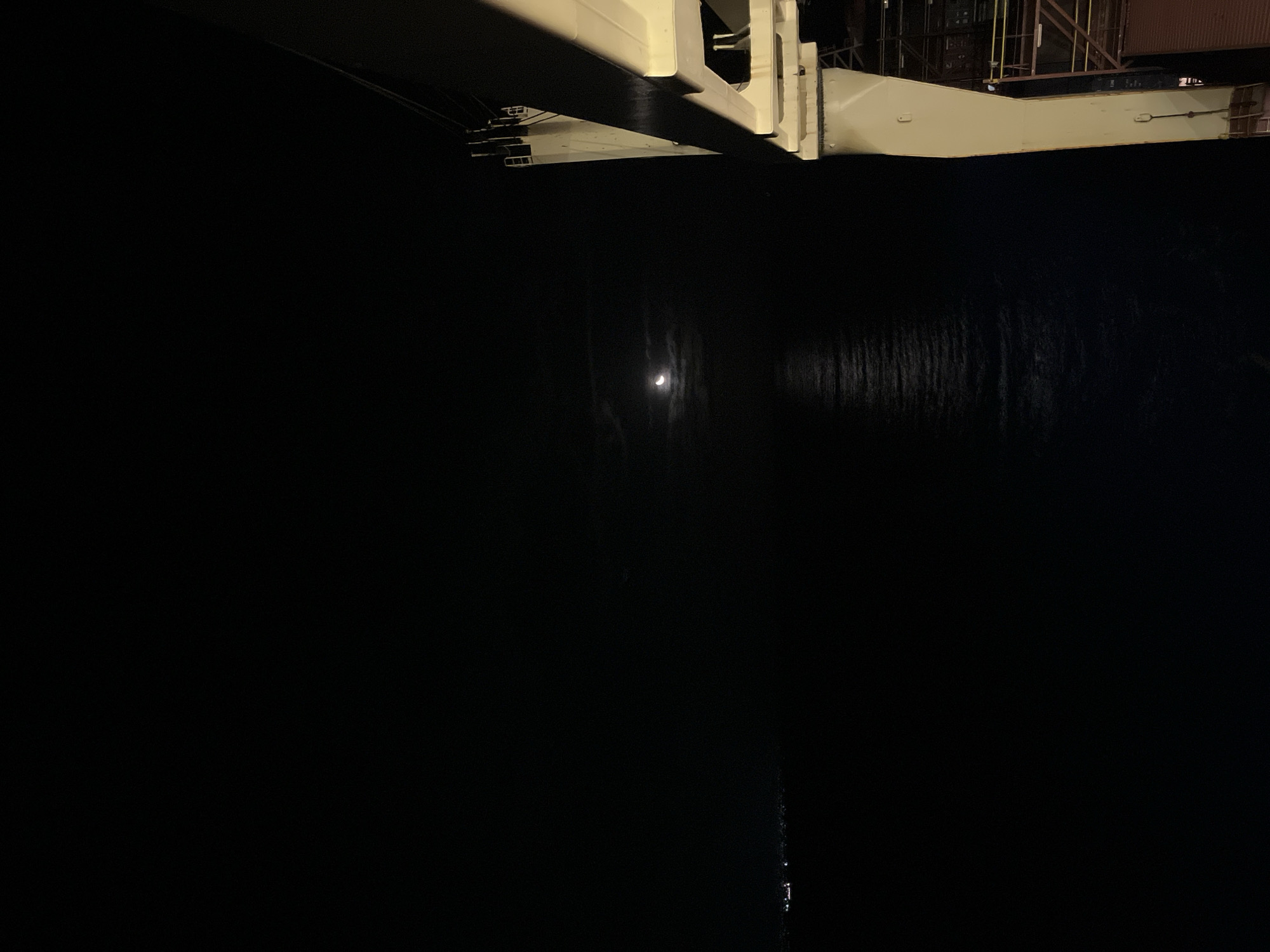 The image depicts a dark, likely nighttime scene. In the upper portion, there's a section of what appears to be a white or light-colored metal structure, potentially a part of a building or industrial machinery. Below this structure, the majority of the frame is filled with dark water, reflecting a crescent moon or bright light. The surrounding environment is not clearly visible, but it suggests a waterfront or dock area, given the presence of water and the industrial-like structure. The city and specific location are not identifiable based on the given visual information.