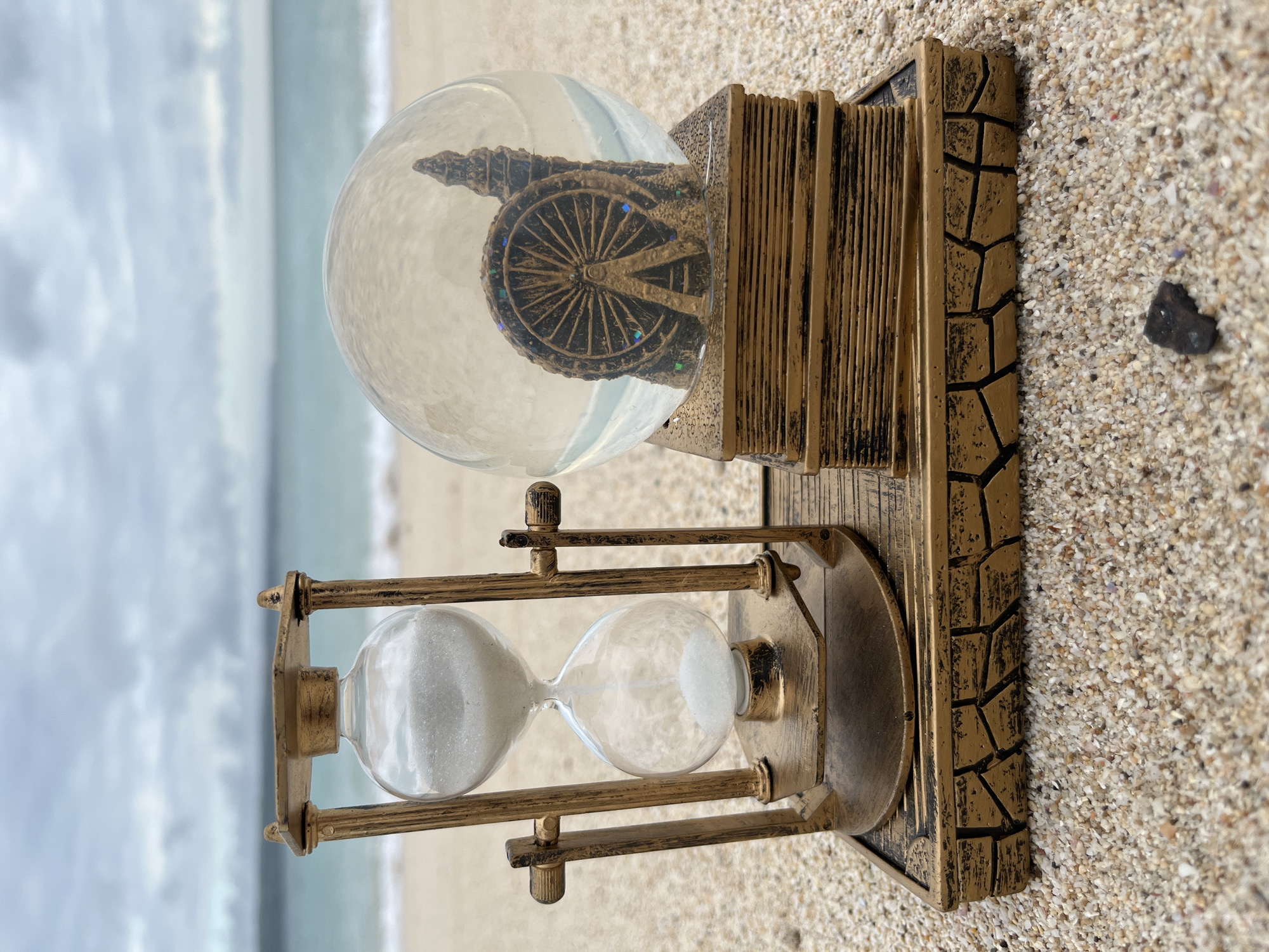 In Kuta, Indonesia, an hourglass sits on a sandy beach next to a snow globe. The hourglass, made of golden metal and glass, contains white sand, with most of it accumulated in the bottom bulb. To its right, a golden snow globe, shaped like a stack of books, encloses a miniature ferris wheel and a tall building. The snow globe sits on a base that resembles a cracked stone pattern. The beach is covered in small pebbles and sand. In the background, the ocean meets a cloudy sky.