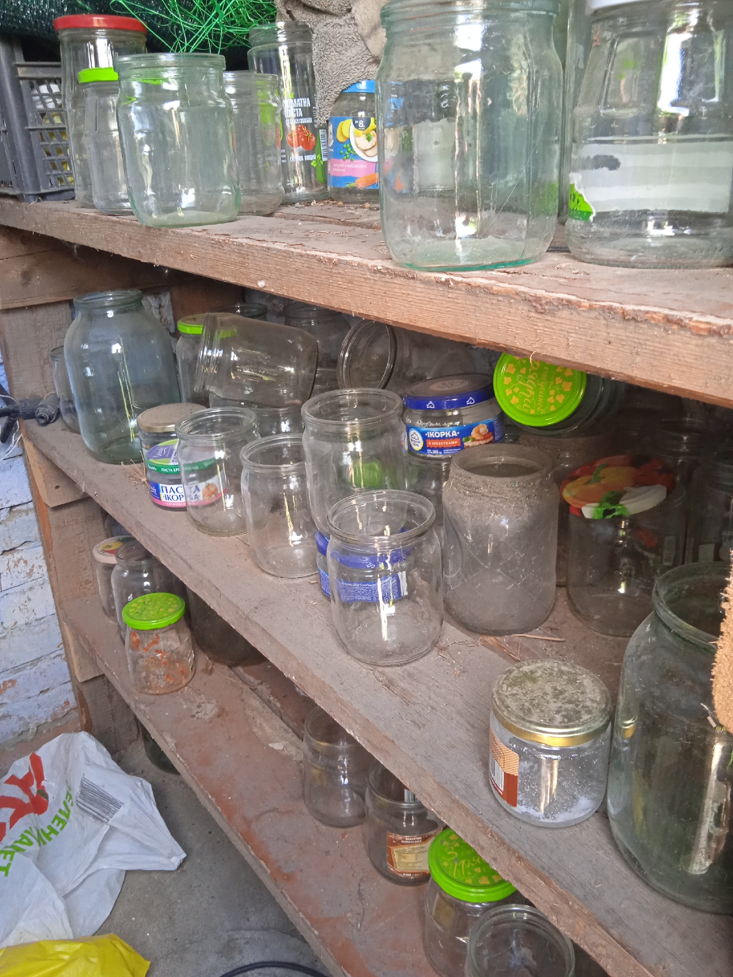The image shows a cluttered storage shelf with numerous glass jars of various sizes. The shelf appears to be made of unfinished wood and is mounted on a wooden structure. In the background, a brick wall is partially visible on the left. On the floor below the shelf, a white plastic bag and a yellow object are present. The jars are mostly empty, some with lids, and a few appear to contain remnants of food items or other substances. The lighting suggests an indoor or shaded outdoor environment. The overall scene depicts a storage area likely used for preserving food or collecting recyclables.
