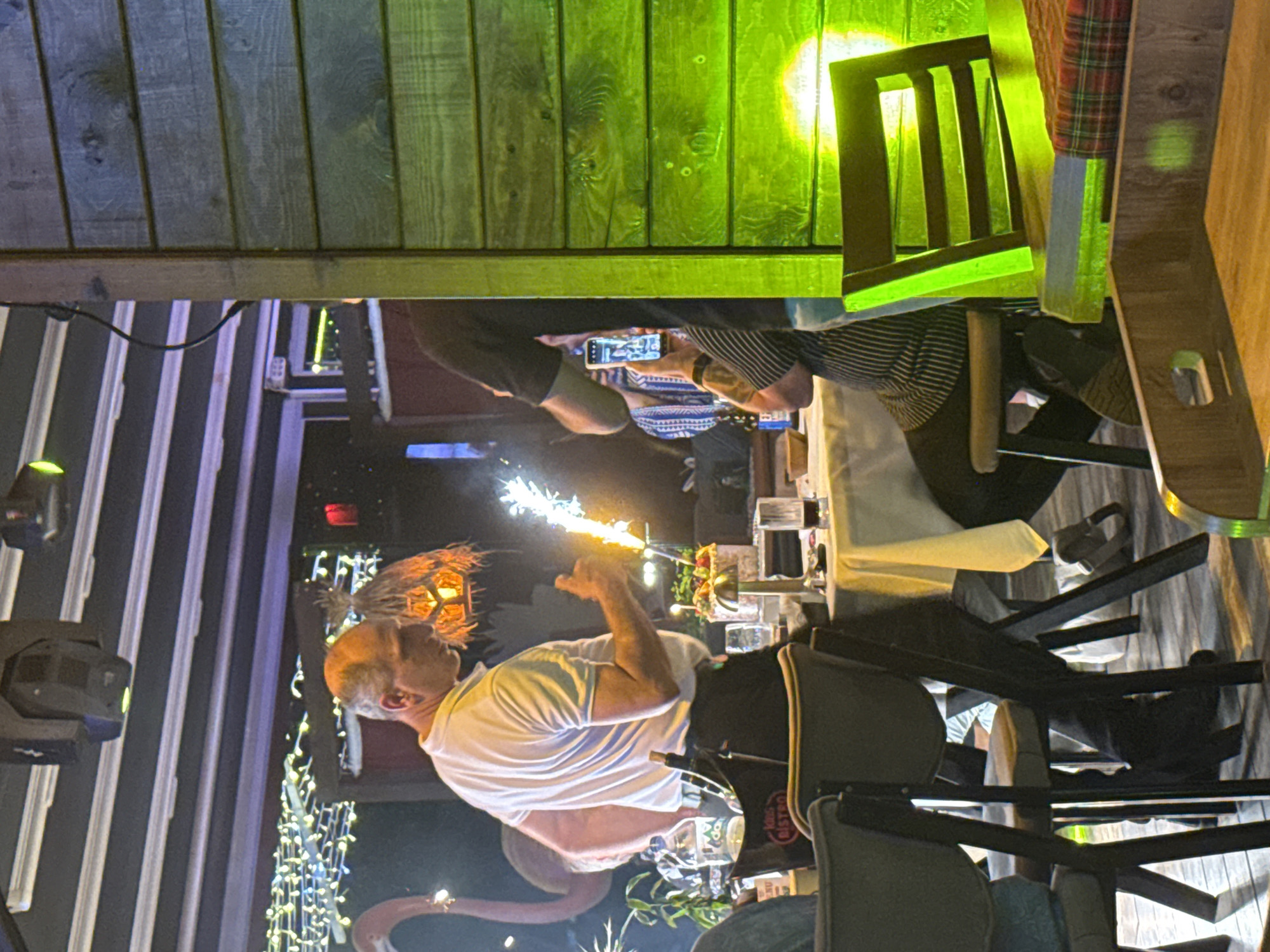 The image captures a lively evening scene, likely in a restaurant or bar. The central figure is a man with light hair, wearing a white t-shirt, who is holding a sparkler that emits bright, fiery sparks. His face is partially visible as he looks towards the sparkler.
In the background, another person, wearing a striped shirt, is seated at a table and is actively using a smartphone, presumably to photograph or video record the event. Parts of other individuals are also visible, suggesting a gathering.
The setting features multiple tables, some with white tablecloths, and chairs, along with various glassware. A bottle on one table clearly displays the text "BISTRO" on its label. Decorative elements include strings of fairy lights, a flamingo-shaped decoration, and what appears to be a tiki torch-style light fixture. Overhead, the wooden ceiling is bathed in a distinct green light, adding to the festive atmosphere, complemented by spotlight-like lighting fixtures on the left. The scene suggests a celebration or special occasion taking place in the evening or night.