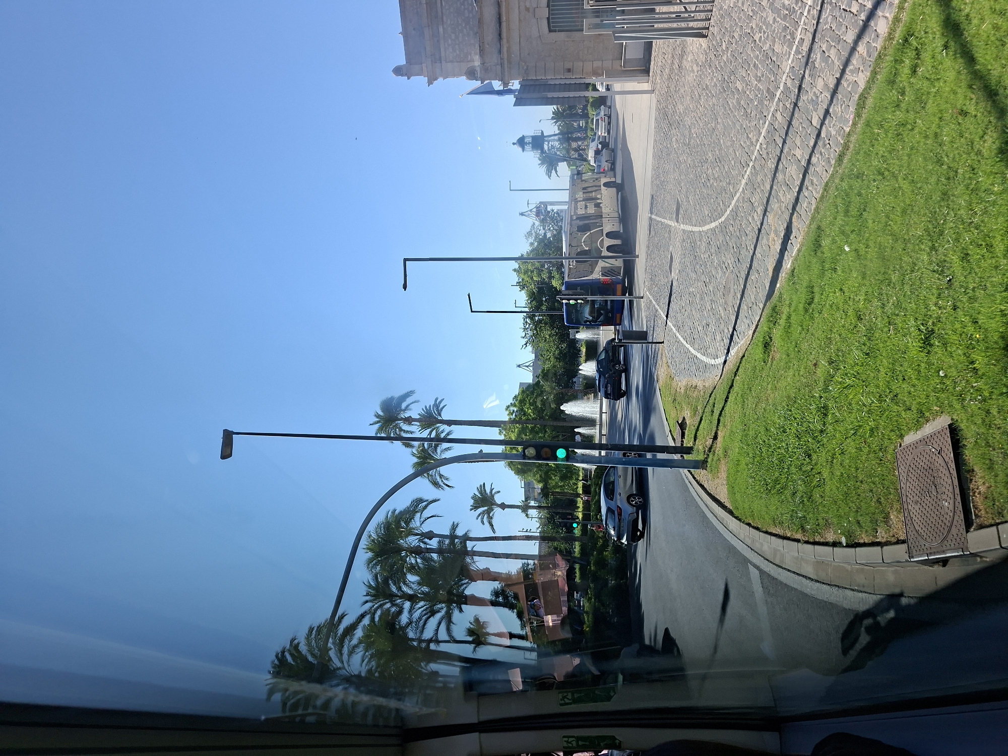 The image depicts a street scene in Alicante, Spain, as viewed from inside a vehicle. The foreground features a patch of green grass and a cobblestone sidewalk. A dark grey car is visible on the asphalt road to the left, followed by a line of palm trees. Further down the road, a blue and white bus with the text "alicante airport" is traveling. A traffic light displays a green signal. In the background, a large building with a facade resembling stone is situated on the right side of the image. The sky is clear and blue.