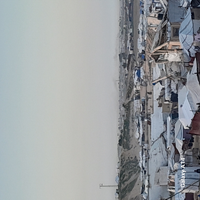 A dense settlement of makeshift dwellings under a pale sky. Predominantly white tents and structures cluster together, some damaged. A dirt path cuts through the camp. In the background, a distant hazy cityscape appears. An antenna is visible on the left.