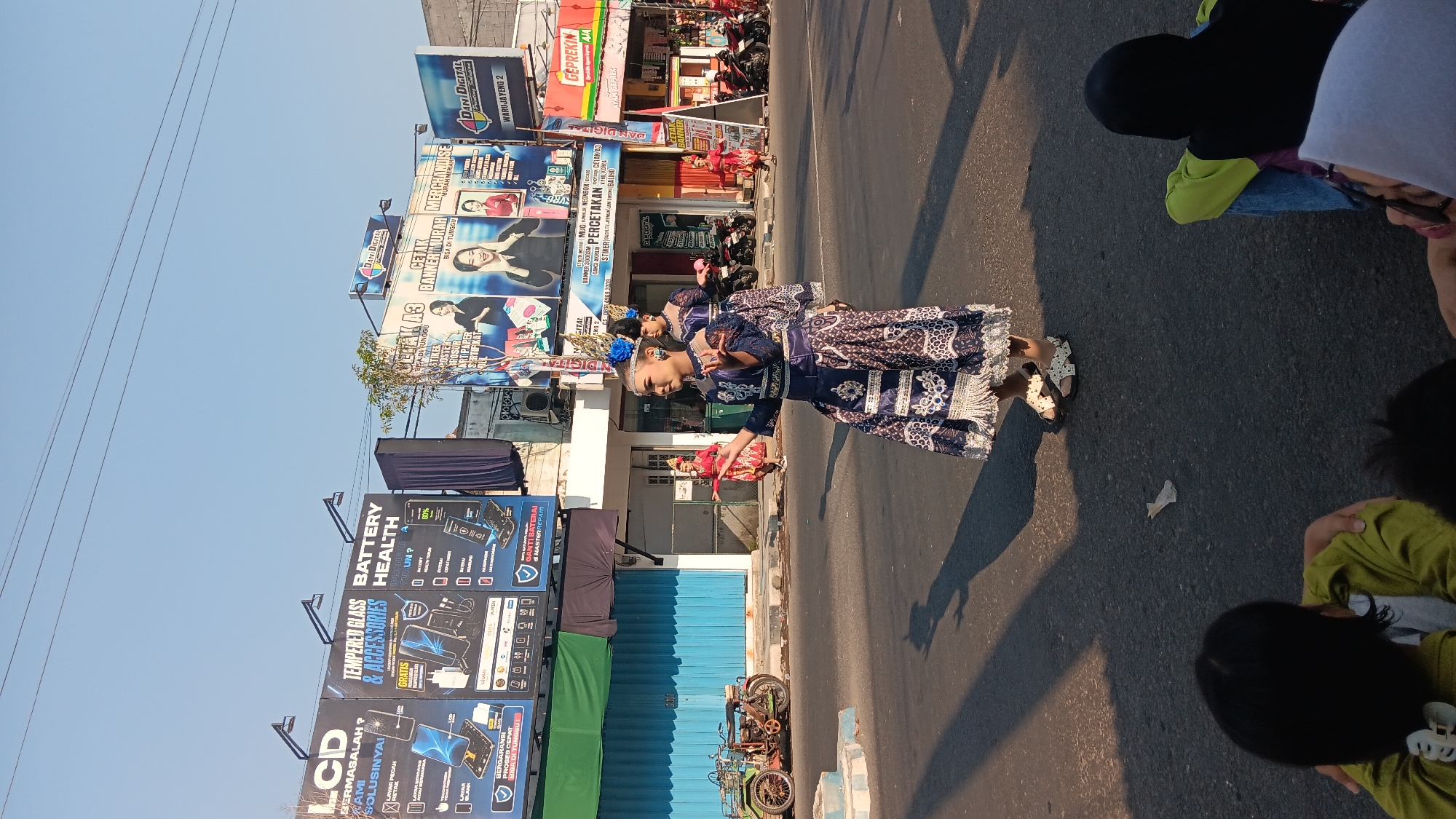 In Warujayeng, Indonesia, an outdoor daytime scene unfolds on a paved street, likely during a parade or performance. A young female dancer, adorned in an elaborate blue and white traditional costume with intricate patterns and a feathered headpiece, is the central figure, captured mid-movement with her arms outstretched. Behind her, another dancer in a similar, dark blue costume, also wearing a headpiece, is visible. Further back, a group of people are observing the performance. To the left, a vintage green motorcycle with a cargo rack is parked on the side of the road, near a building with blue metal roll-up doors. The background features a row of commercial buildings with large billboards advertising services such as "LCD TEMPERED GLASS & ACCESSORIES," "BATTERY HEALTH," "CETAK A3," and "DANI DIGITAL." The sky above is clear and blue, indicating sunny weather, and the ground is marked with shadows. The overall impression is a vibrant cultural event taking place in a commercial district.