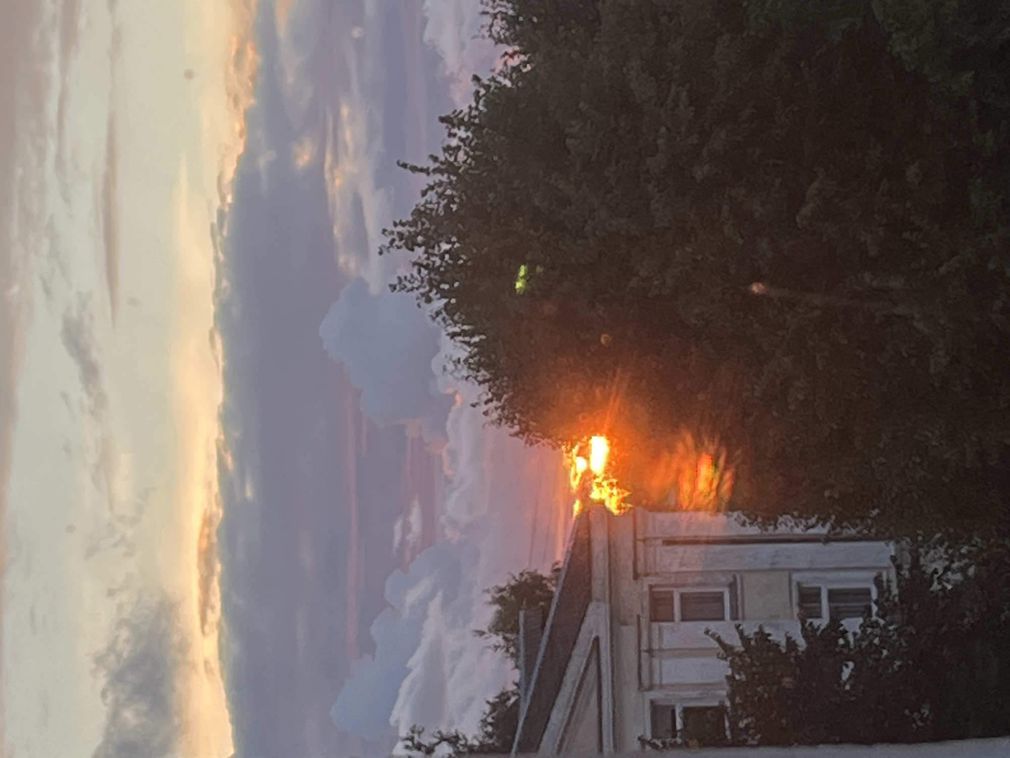 The image captures a sunset viewed from an urban setting in Kohtla-Järve linn, Estonia. The sky is filled with hues of orange, pink, and purple, with scattered clouds. In the foreground, a portion of a building with white columns and windows is visible on the left. To the right of the building, dense green foliage from trees obscures much of the view. The setting sun, a bright orange orb, is partially hidden behind the trees and creates a strong glare, illuminating the leaves and casting warm light. The overall scene suggests a residential or administrative area within the city, characterized by a mix of architecture and natural elements.