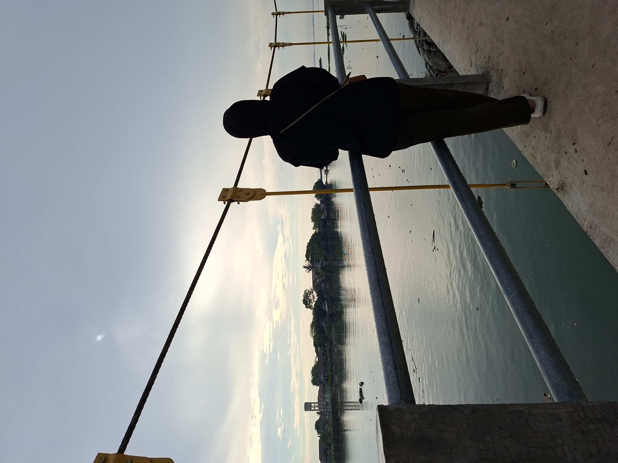 The image shows a person wearing a black hijab and a black hoodie, standing on a concrete walkway next to a body of water. The person is facing away from the camera, looking out at the water and the distant shore. The walkway has a metal railing. In the distance, there are buildings and trees on the opposite bank of the water. The sky is overcast, with some clouds visible. The image appears to have been taken during the day. The location is the Lhokseumawe Reservoir, a man-made lake in Lhokseumawe, Indonesia, which serves as a source of freshwater for the city and surrounding areas.