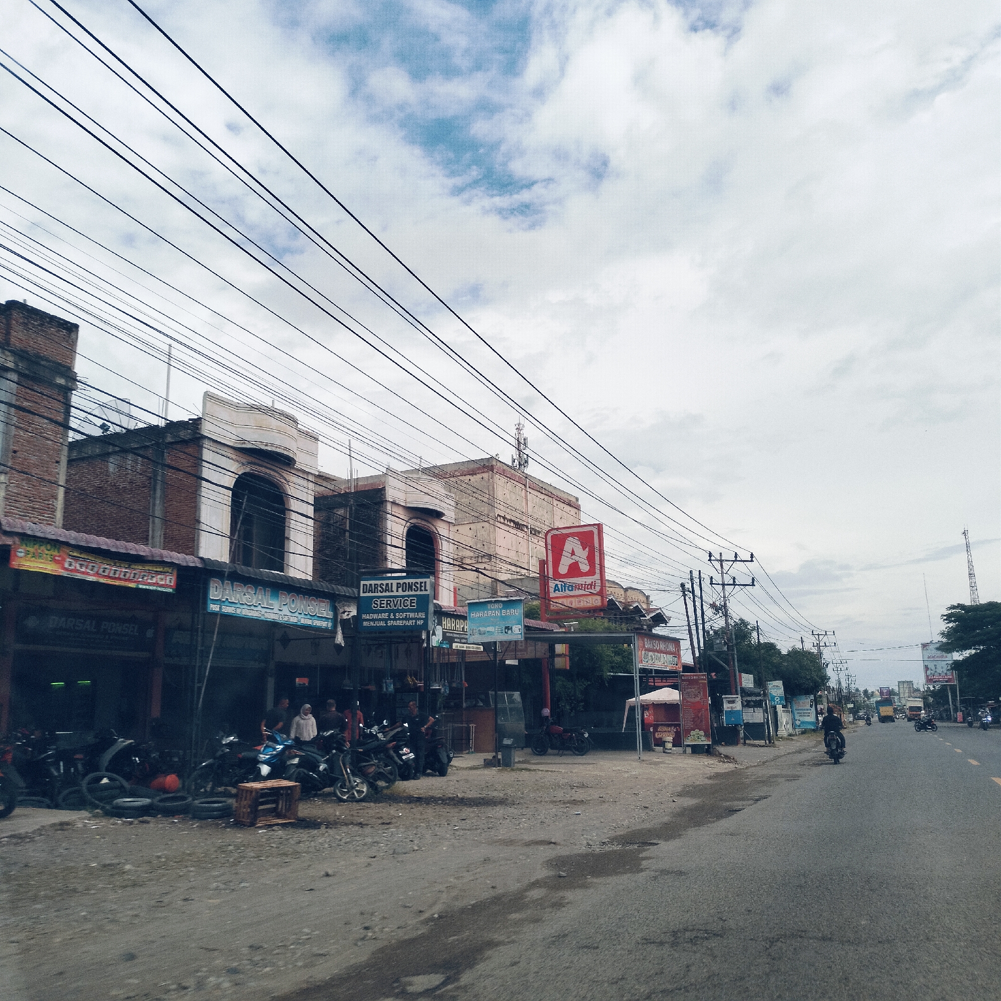 This image depicts a bustling street scene in Desa Meunasah Blang, Indonesia, under a cloudy, overcast sky. The foreground features a dirt and gravel roadside bordering a paved road, which stretches into the distance.
On the left side of the street, a row of commercial buildings houses various shops. Numerous motorcycles are parked along the roadside in front of these establishments, with several people gathered nearby, some standing or sitting. A wooden crate and a pile of tires are visible on the ground, suggesting activity related to vehicle maintenance or sales.
Prominent signs identify several businesses: "DARSAL PONSEL," which is advertised multiple times and specifies "SERVICE," "HADWARE & SOFTWARE," and "MENJUAL SPAREPART HP." Other signs include "TOKO HARAPAN BARU," a large red "A" logo for "Alfamidi," and "BAKSO RHONA" further down the street.
Overhead, a complex web of black utility wires crisscrosses the sky, connected to numerous utility poles that line the road. In the distance, a person is seen riding a motorcycle away from the viewer on the paved road. The background shows more buildings, trees, and a tall cellular tower. The scene conveys a typical daytime atmosphere of commerce and daily life in a local Indonesian town, with the overcast weather providing diffused lighting.