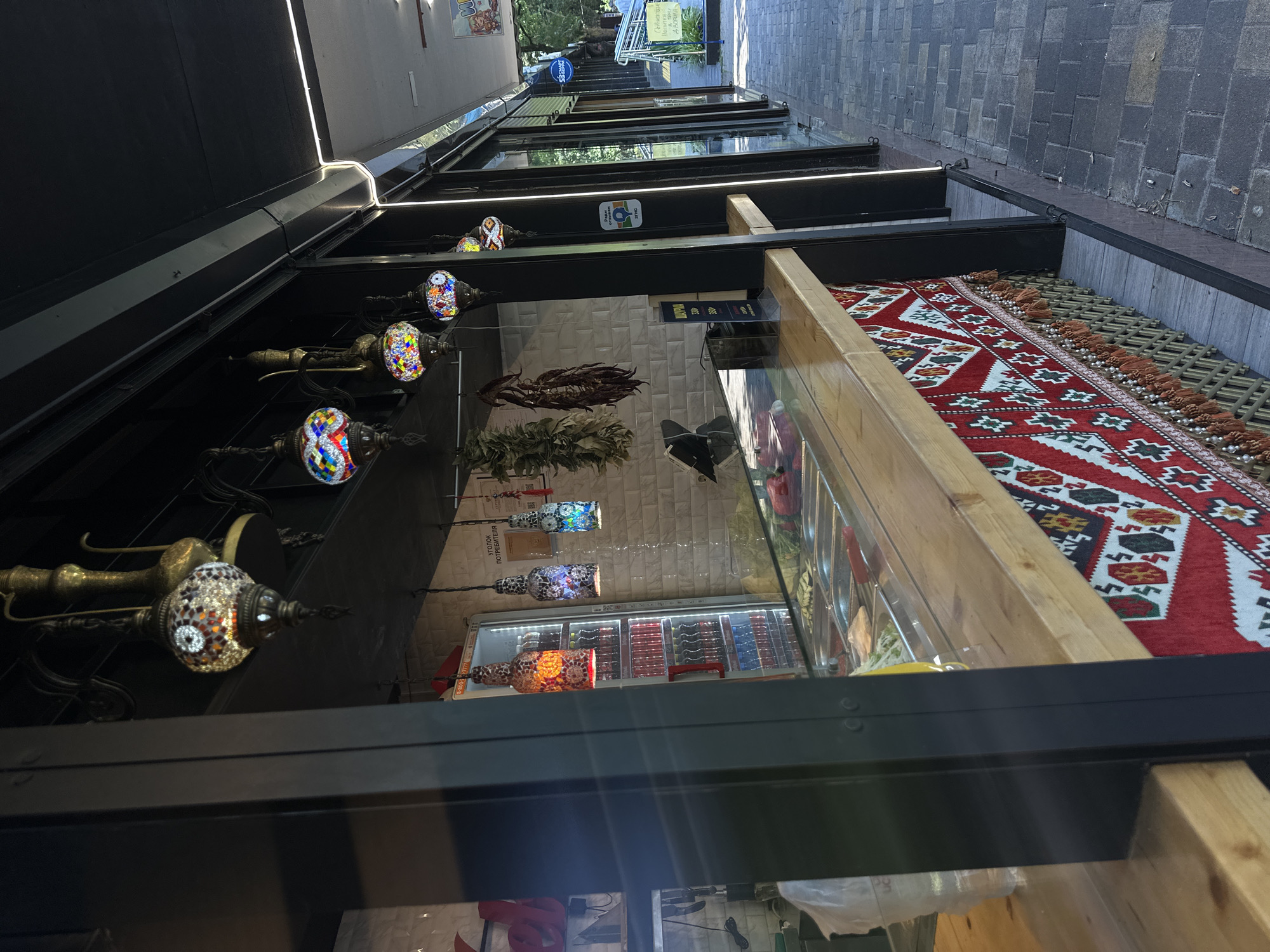 The image depicts an outdoor food stall located in the city of Sochi, Russia. The stall is characterized by its decorative elements, including mosaic lamps hanging from the ceiling and a patterned textile draped over the front counter. Behind the counter, a refrigerated display case is filled with beverages, and a variety of ingredients are visible in stainless steel trays, suggesting food preparation. The stall appears to be open for business, with a clean and organized setup. The surrounding environment features a brick-paved walkway and other adjacent stalls or establishments, indicating a commercial area. The overall impression is one of a typical street food vendor in a bustling urban setting.