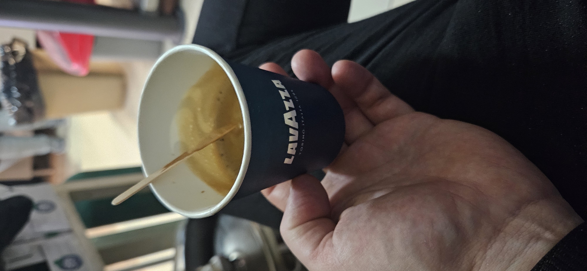 A person is holding a blue Lavazza paper cup filled with coffee. A small wooden stir stick is partially submerged in the liquid. The person's hand is in the foreground, and dark clothing is visible behind it. A blurred background reveals furniture and various objects, including what appears to be a red container. The exact location within Sofia, Bulgaria, cannot be determined from the image.