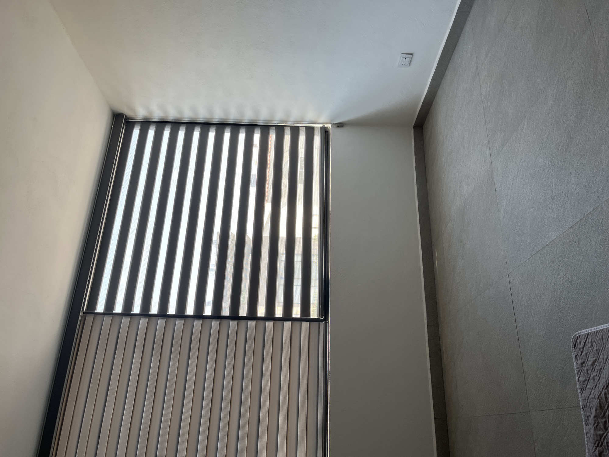 The image depicts an interior space in Puebla, Mexico, featuring a window with vertical blinds, part of which is open, revealing the exterior view of buildings. The interior is characterized by a neutral color palette with a white ceiling and light gray walls, one of which is textured. The blinds are black and white, and a lower section of the window also has patterned vertical blinds. The architectural details are modern, suggesting a residential or commercial building.