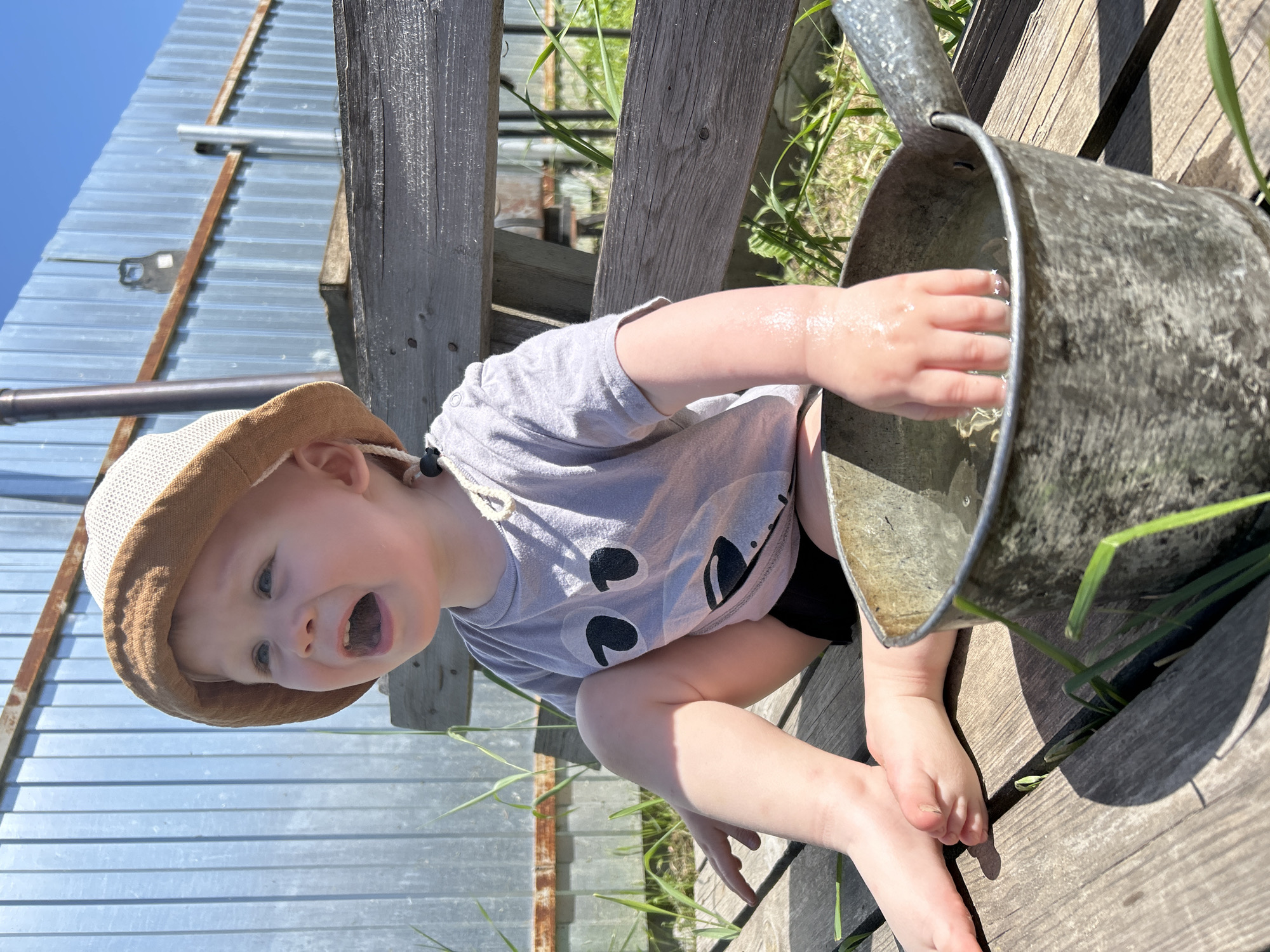 A young child with a light complexion is playing outdoors with water in Chaikovsky, Russia. The child, who is wearing a gray T-shirt with dark round eyes printed on the front, black shorts, and a tan hat, is seated on a weathered wooden surface, possibly a deck or a raised platform. The child's right hand is submerged in the water inside a large, galvanized metal bucket, while their left hand rests on the wooden surface beside them. The child is looking upwards with an open mouth. Adjacent to the child is a wooden fence, and beyond that is a corrugated metal sheet wall. Patches of grass and vegetation are scattered around. The weather appears sunny.